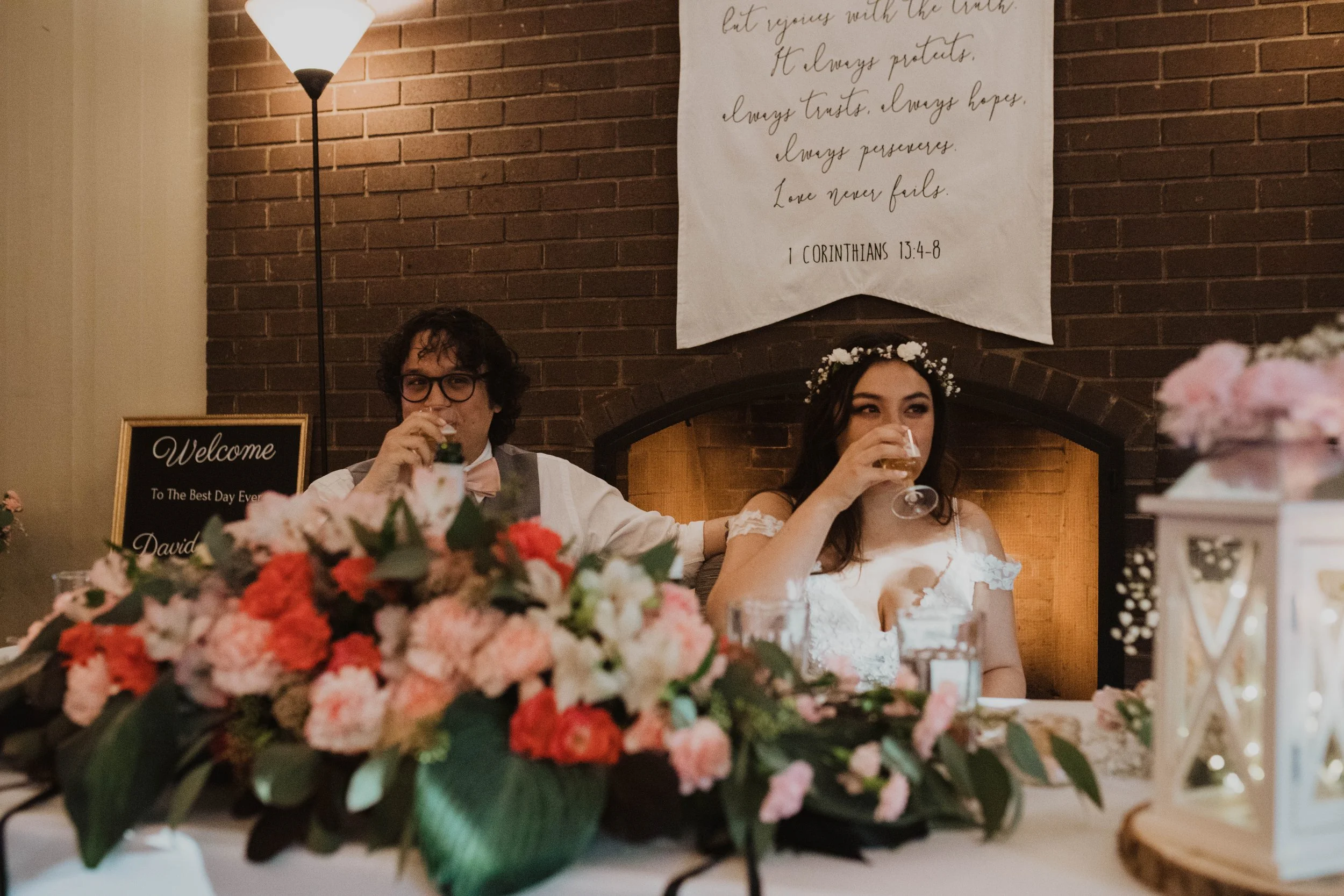 A bride and groom seated at a table during their wedding reception, drinking from glasses, with floral centerpieces in front of them. They are smiling and sitting in front of a brick wall with a hanging banner displaying a Bible verse. There is a lit