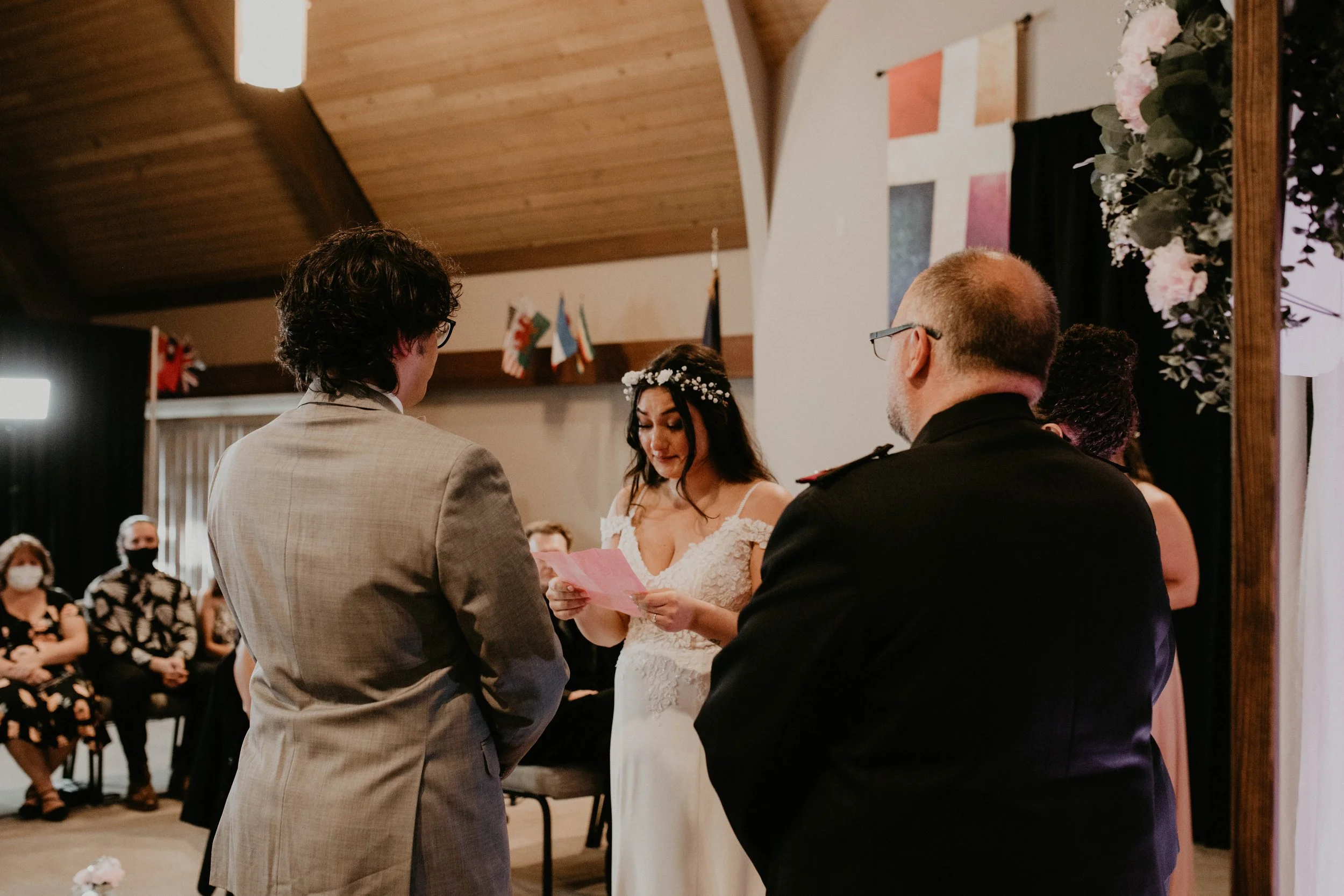 Bride and groom standing during their wedding vows, with officiant reading from a paper, guests seated in the background, some wearing masks, and decorated with flags and floral arrangements. Seattle, WA wedding photography.