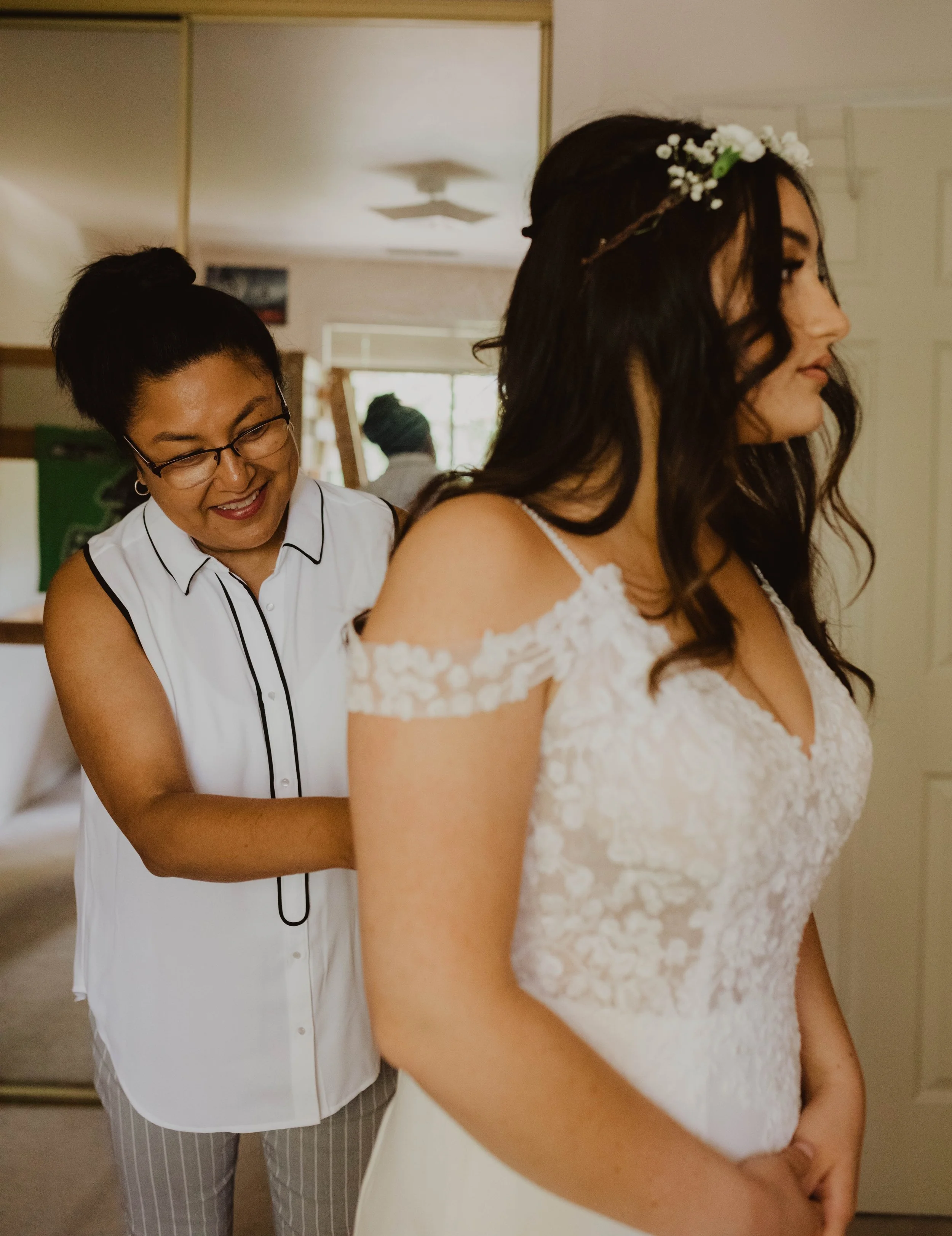 A bride with long dark hair and a floral headband wearing a white lace wedding dress, standing indoors with an open door in the background, while another woman helps her with her dress. Seattle, WA wedding photography.