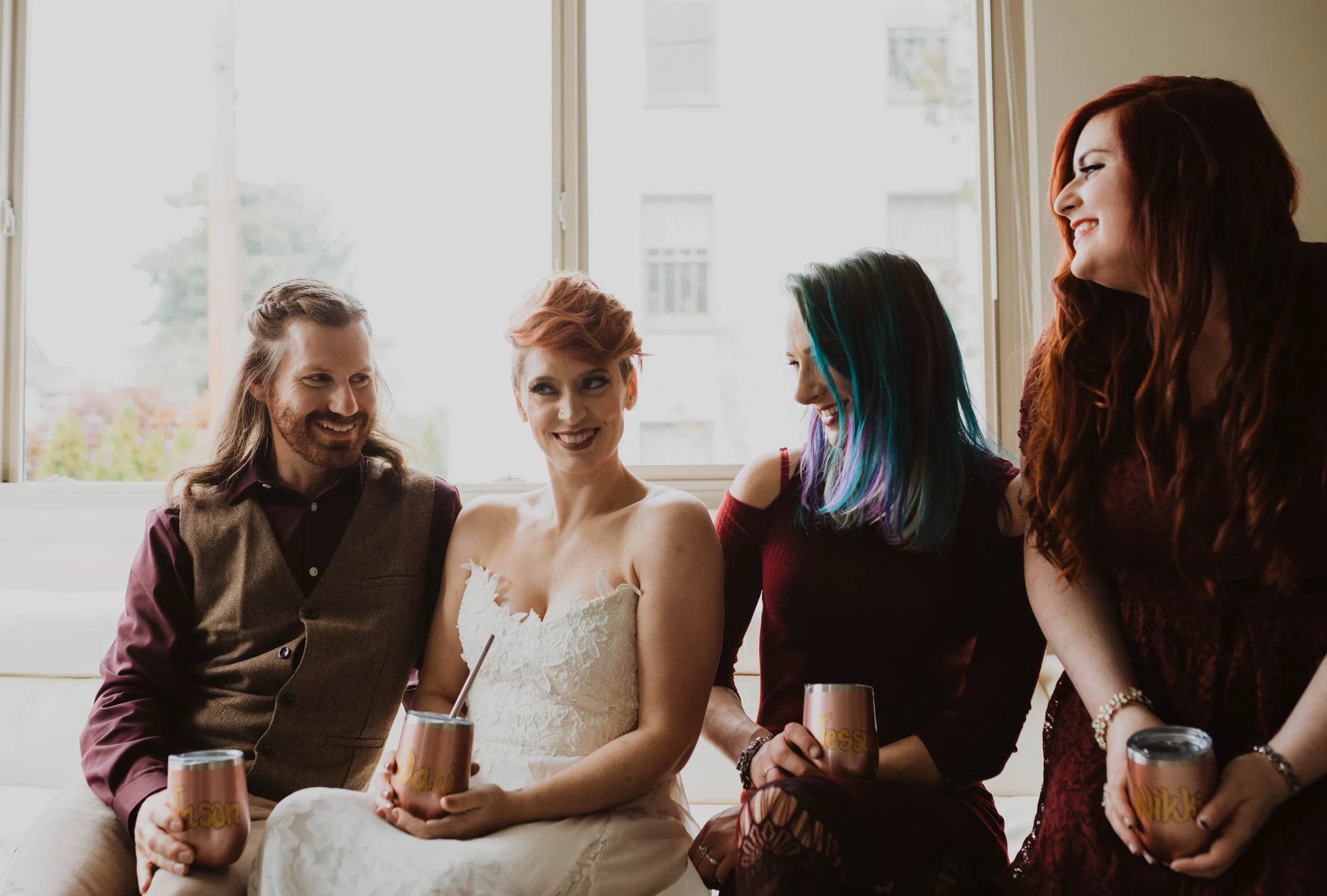 Four friends sitting together on a couch, smiling and holding drink cups, near a large window with trees outside. Pioneer Square, Seattle, WA wedding photography.