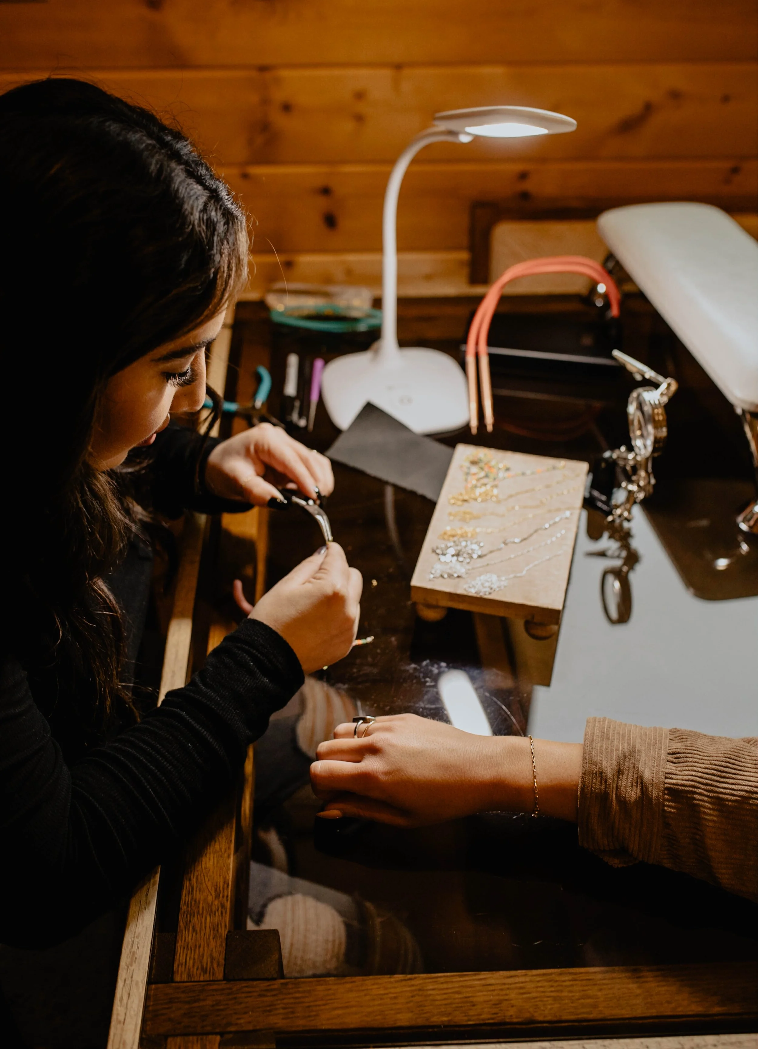 A woman making jewelry at a crafting station with tools, jewelry pieces, and a jewelry display stand, illuminated by a desk lamp, with another person's hand resting on the table. Seattle professional head shot photography