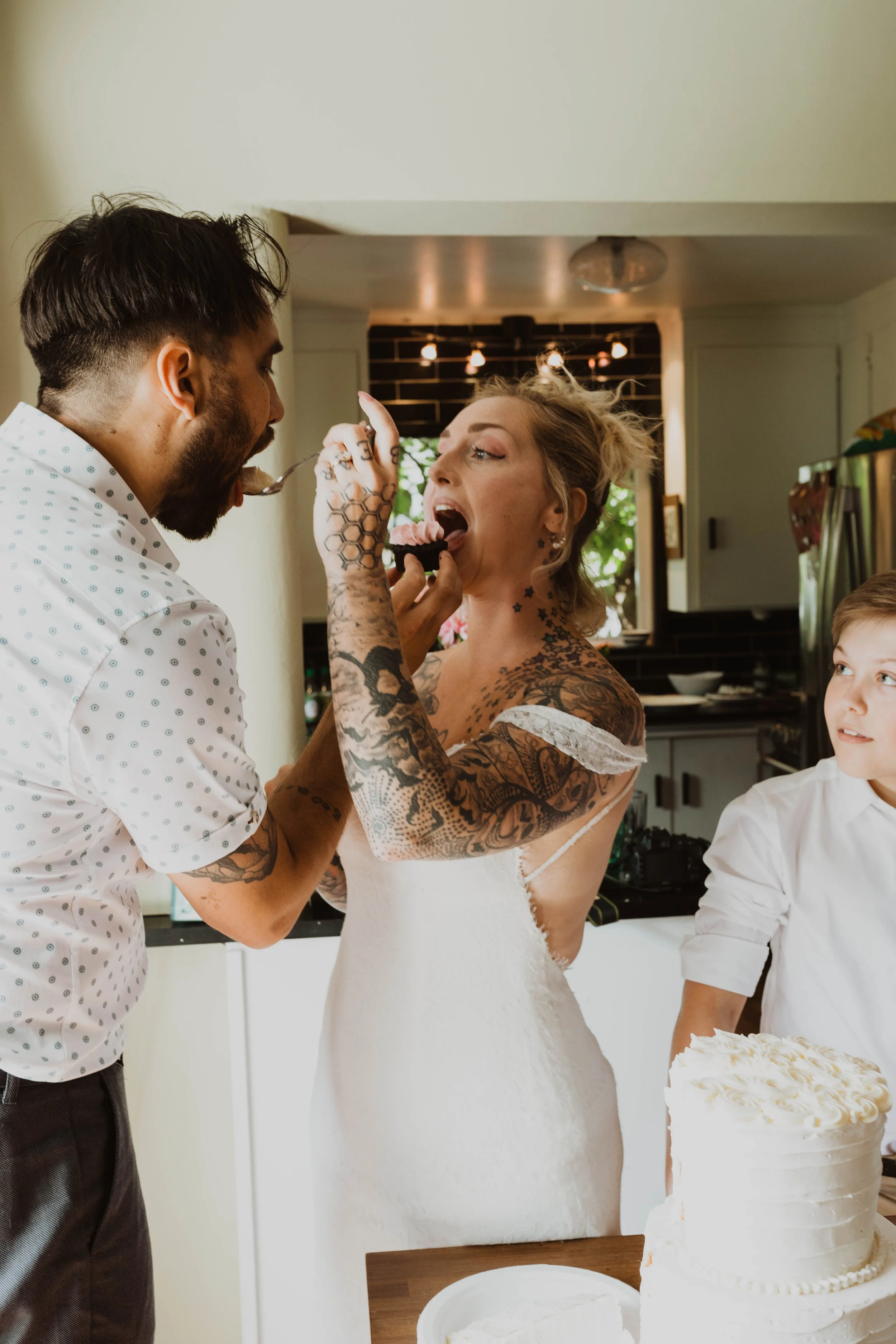A woman in a wedding dress is feeding cake to a man at a celebration, with a young boy watching nearby. Seattle, WA wedding photography.