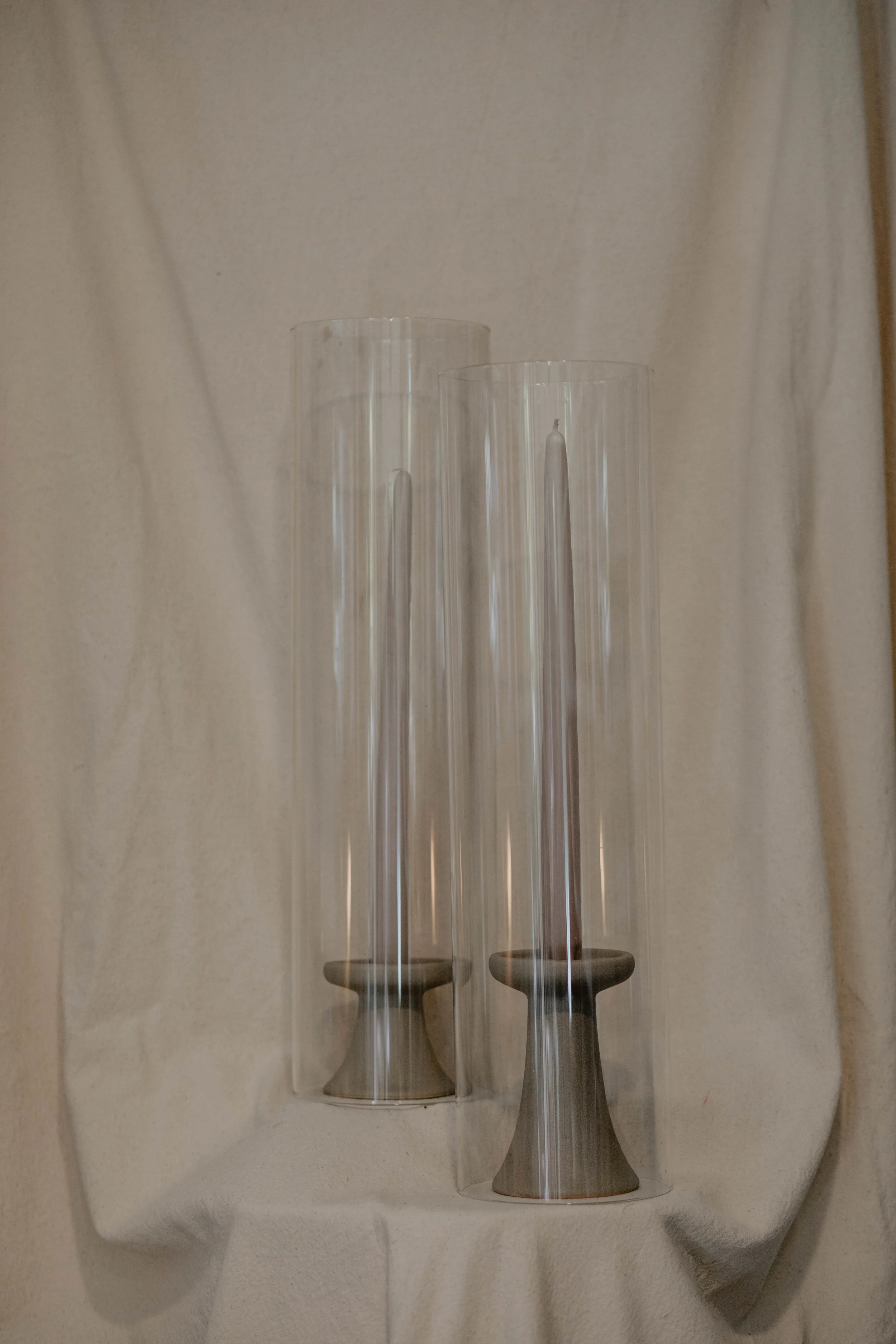 Two modern candle holders with tall, cylindrical glass covers, placed against a beige curtain background.  Seattle product photography
