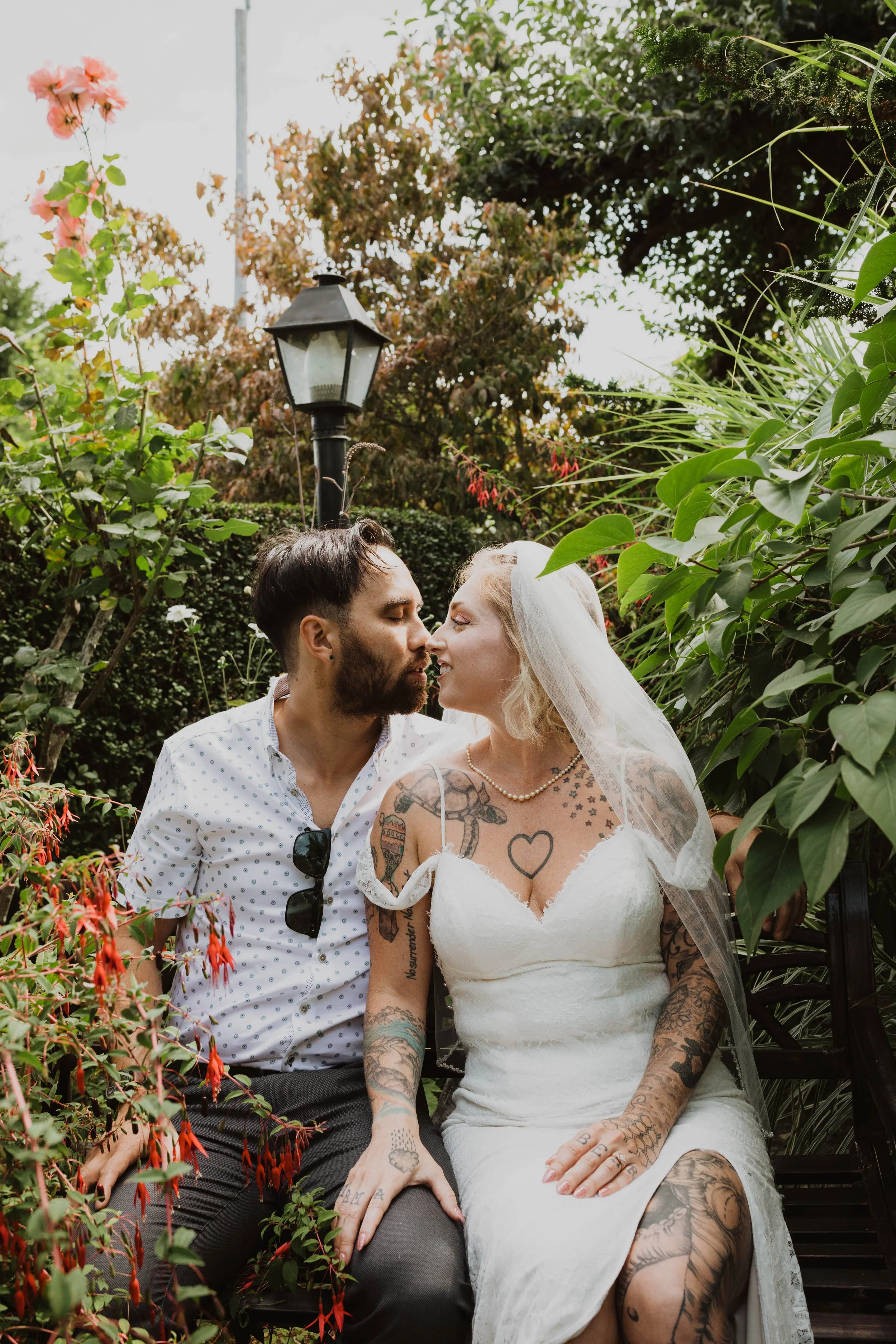 A couple dressed in wedding attire sitting close together on a park bench, surrounded by green plants and with a vintage street lamp in the background, about to kiss. Seattle, WA wedding photography.