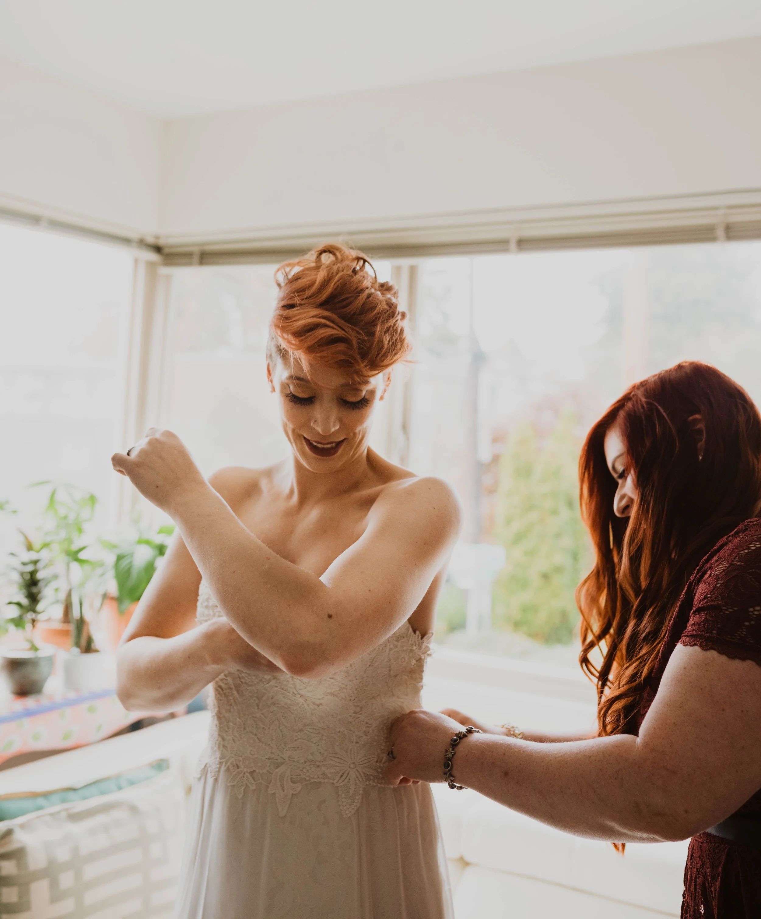 A bride getting ready, assisted by another woman, in a brightly lit room with large windows and potted plants. Pioneer Square, Seattle, WA wedding photography.