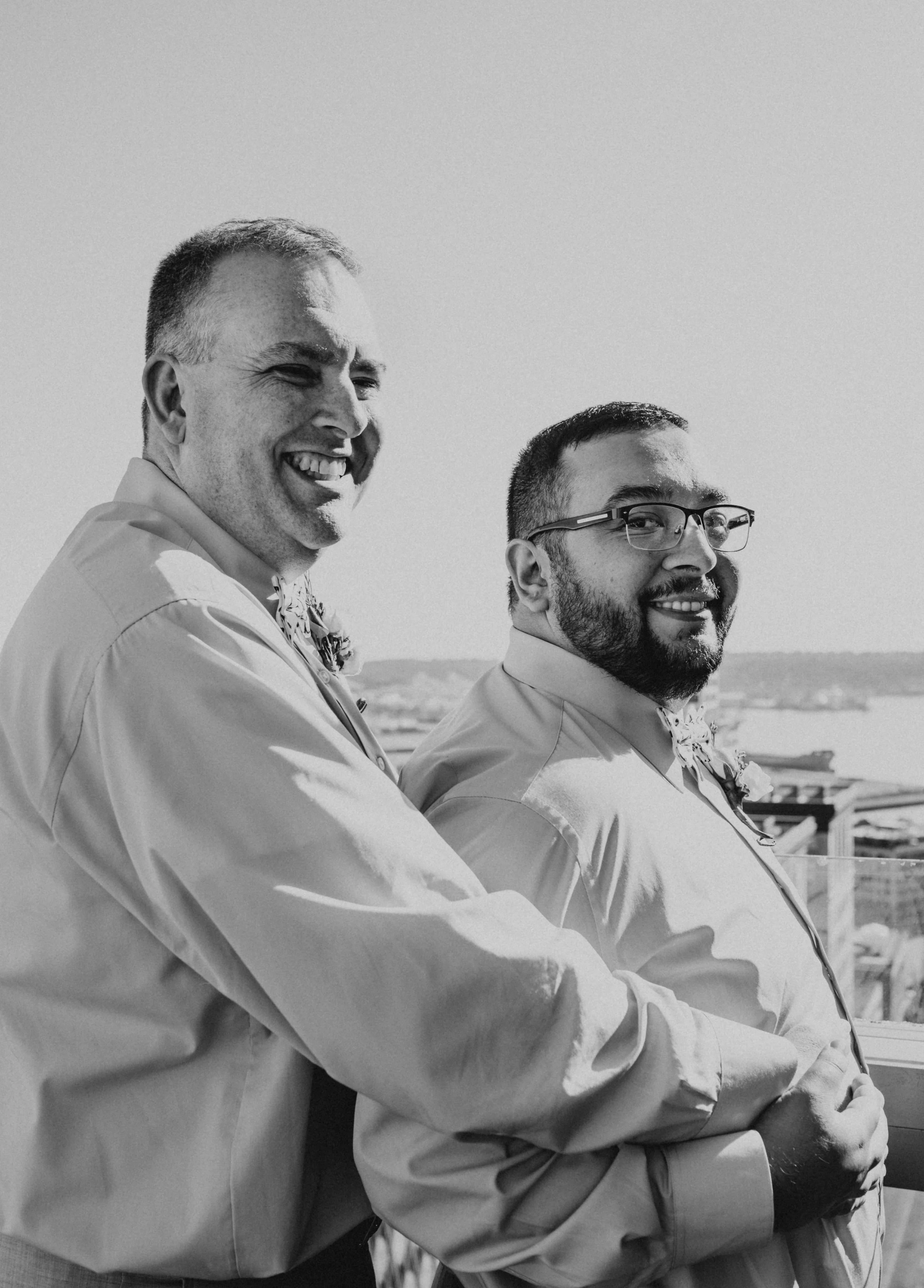 Two smiling men in dress shirts with boutonnieres, standing outdoors with a water body and cityscape in the background. Seattle Municipal Courthouse wedding photography.