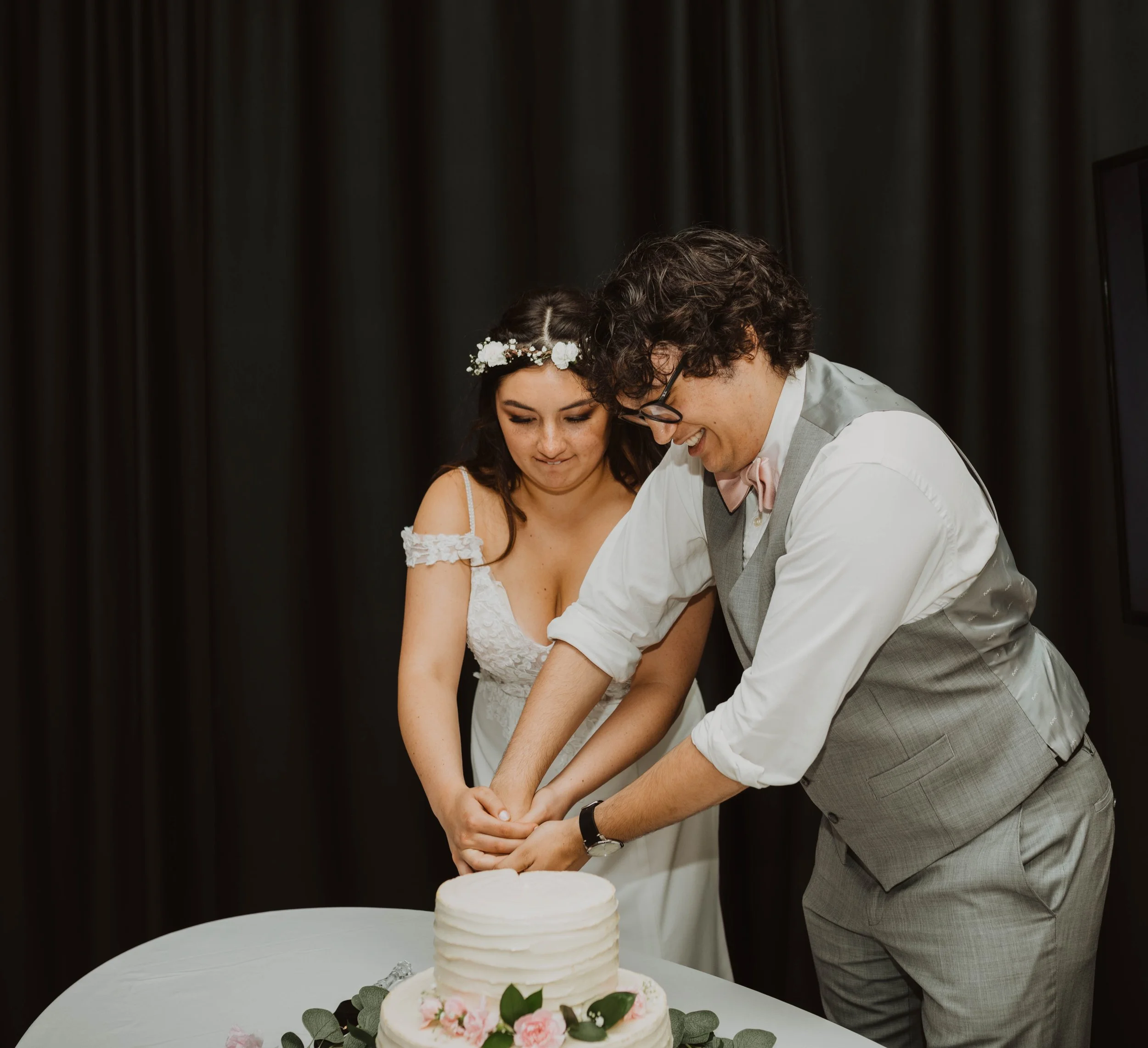 A bride and groom cutting a wedding cake together, both smiling, in front of a dark background. Seattle, WA wedding photography.