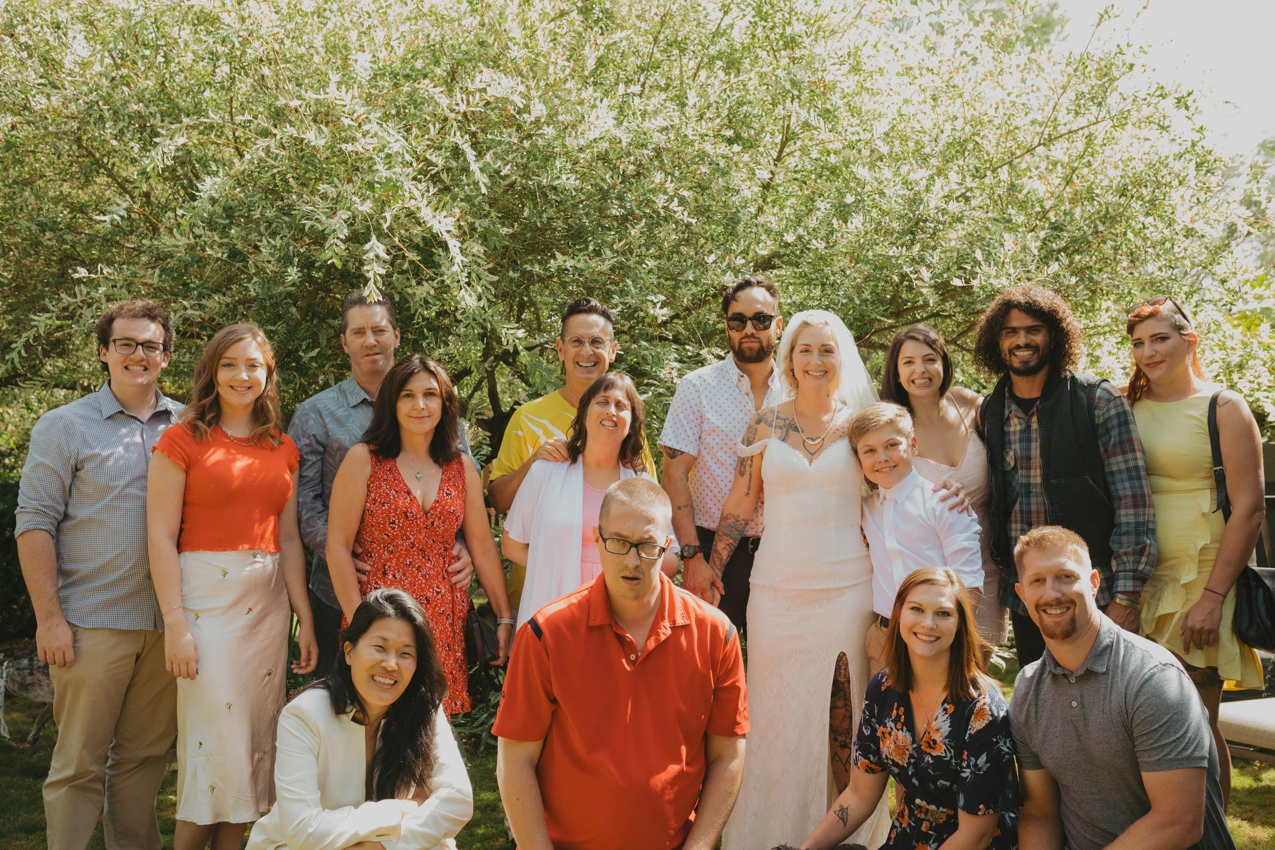 Group of people gathered outdoors in front of a large tree, smiling for a photo, some dressed casually and others in formal attire including a bride in a white wedding dress. Seattle, WA wedding photography.