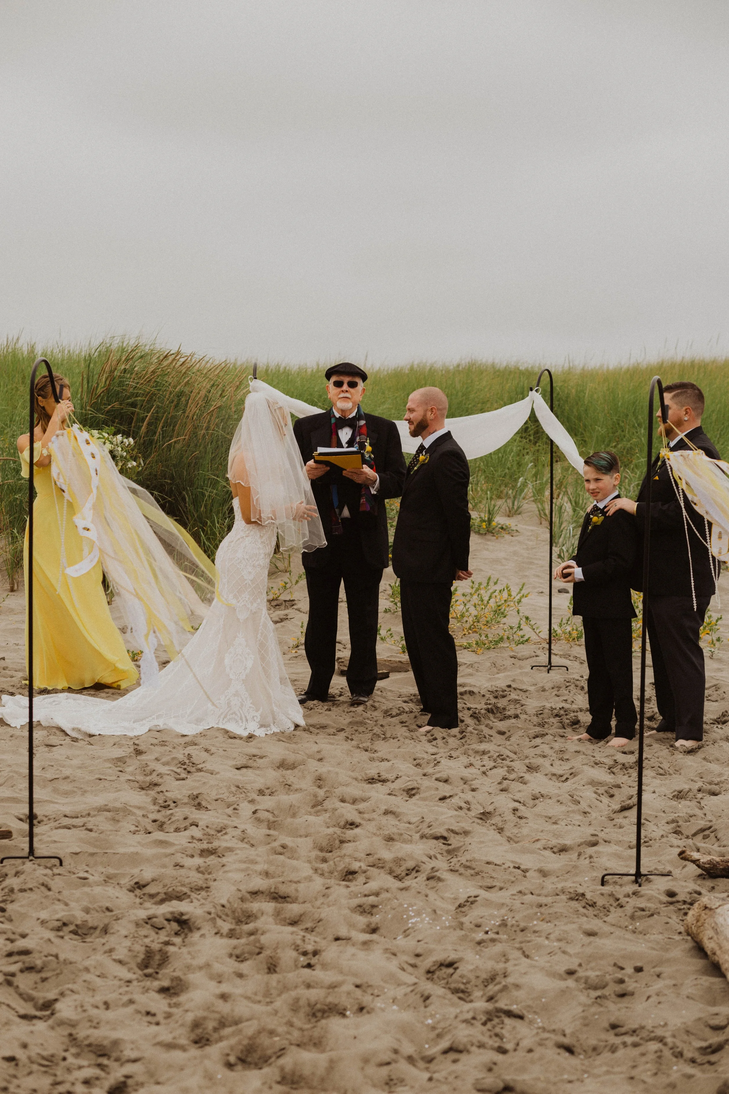 A beach wedding ceremony with a bride in a lace wedding dress and veil, a groom in a black suit, and an officiant in a black jacket and hat, surrounded by bridesmaids in yellow dresses and groomsmen in black suits, on a sandy beach with grass and a c