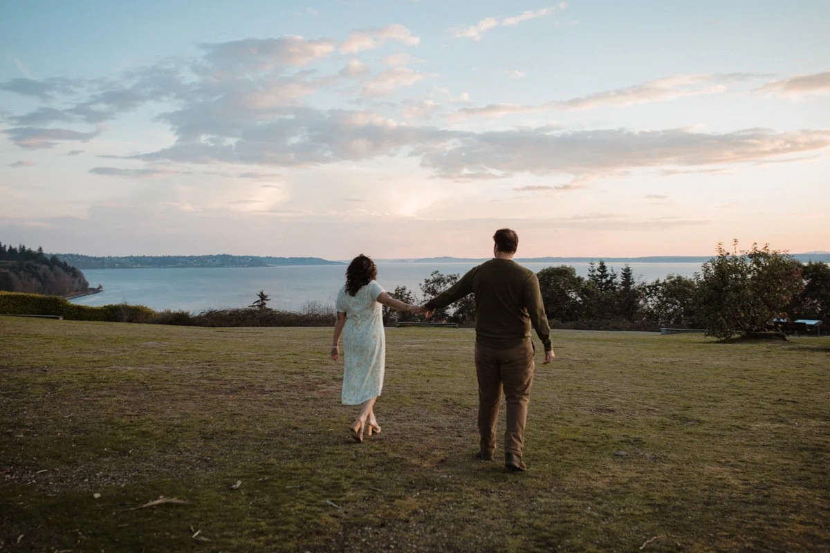 A couple holds hands while walking on a grassy area near the water at sunset, with clouds in the sky and distant trees along the shoreline.