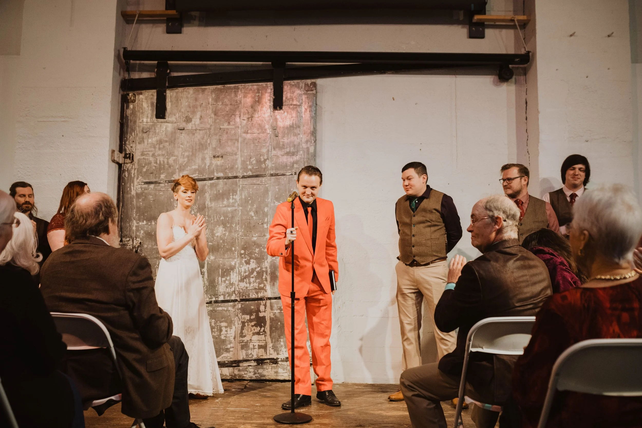 A wedding ceremony with a man in a bright orange suit holding a microphone in front of seated guests, a woman in a white wedding dress beside him, and several men in suits on a stage with a rustic door backdrop.