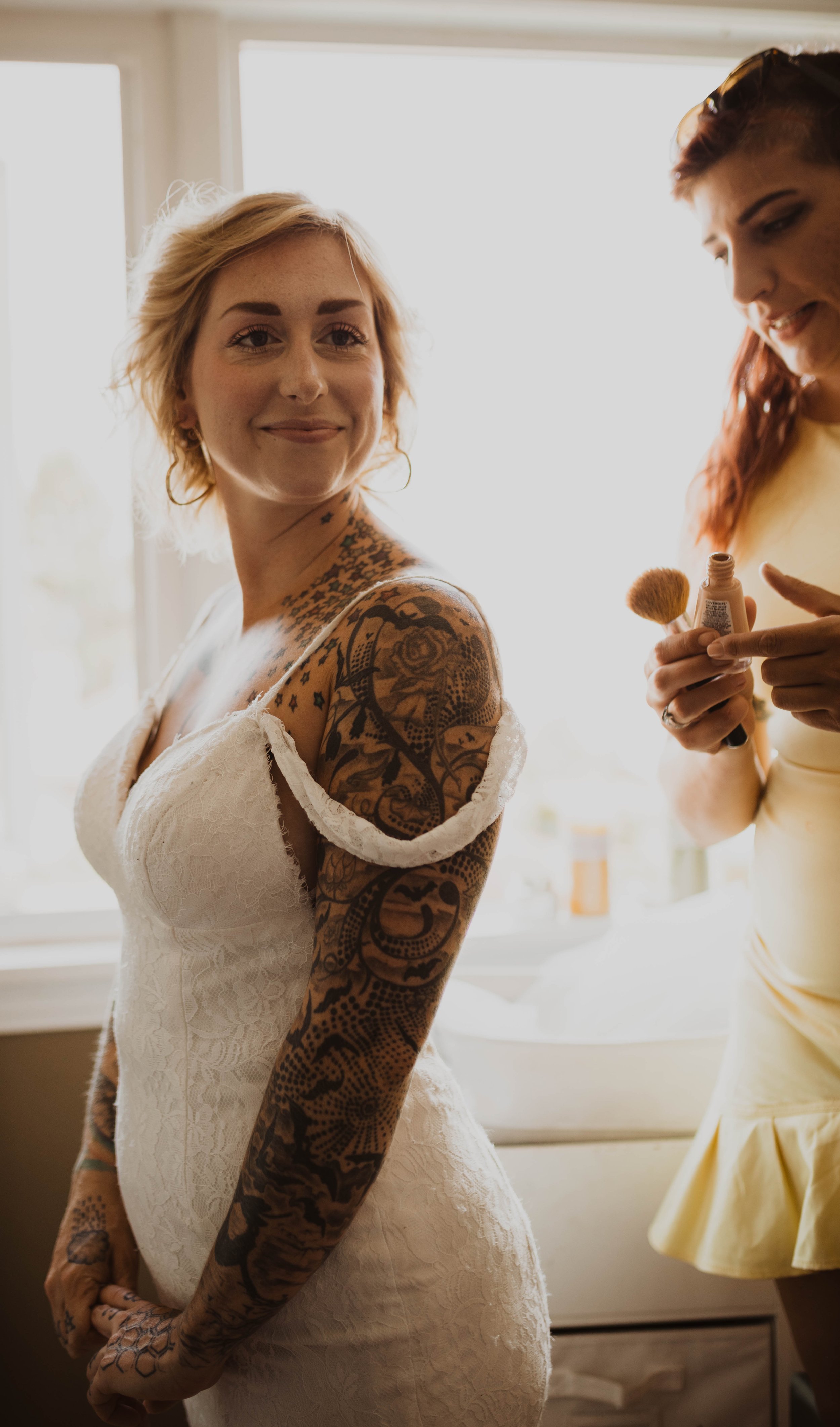 A woman in a white lace dress smiling as she gets her makeup done by another woman. The woman being helped has short blonde hair, tattoos on her arms, and is standing by a window with natural light. Seattle, WA wedding photography.