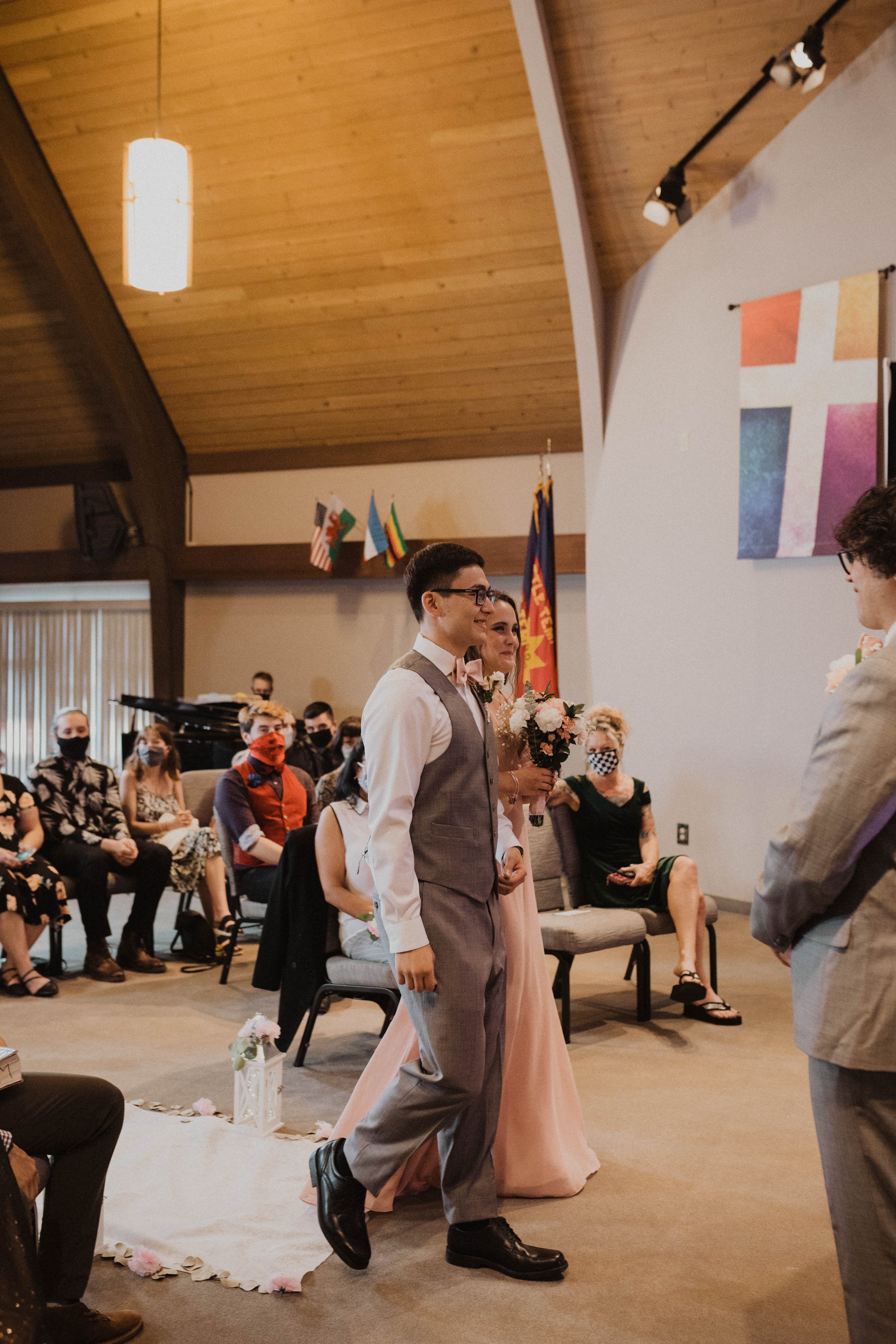 A wedding ceremony with a groom in a gray suit and a bride in a peach dress holding a bouquet. They are standing facing a man in a gray suit. Wedding guests sit in the background, many wearing masks, in a room with a wooden ceiling and flags hung on 