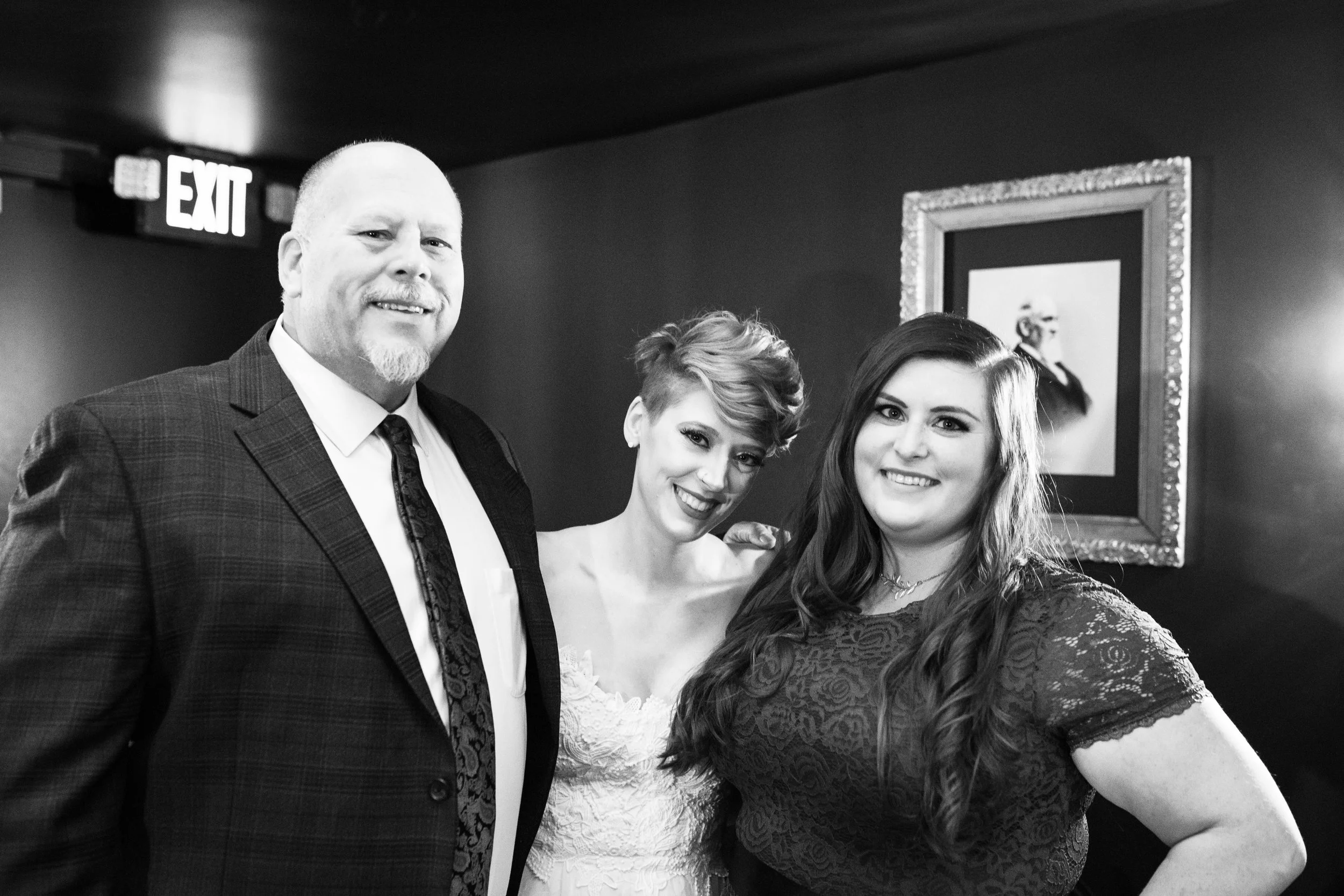 Three people posing together indoors, smiling, with a framed picture on the wall behind them and an exit sign above. Pioneer Square, Seattle, WA wedding photography.