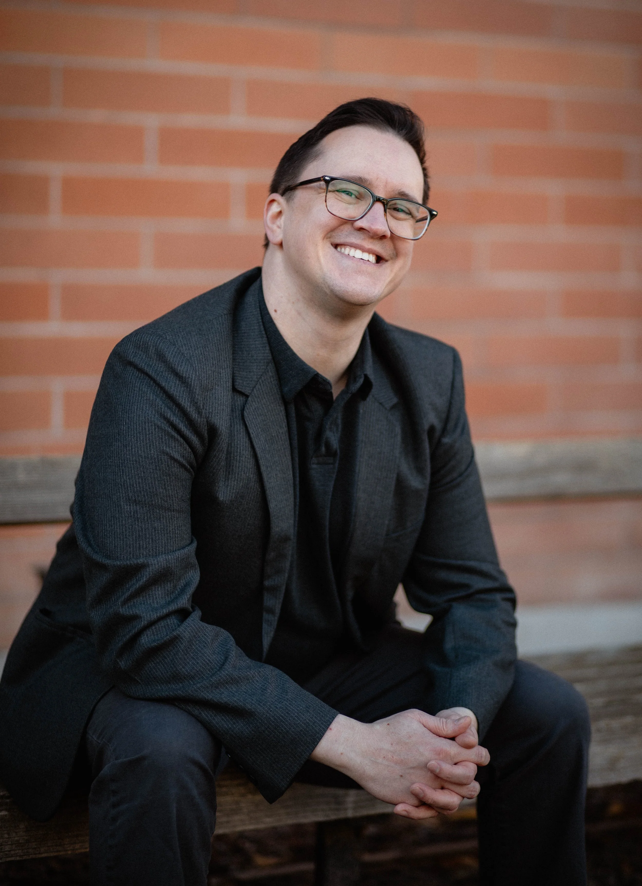 A man with glasses, wearing a black blazer, sitting on a wooden bench in front of a brick wall, smiling at the camera. Seattle professional head shot photography
