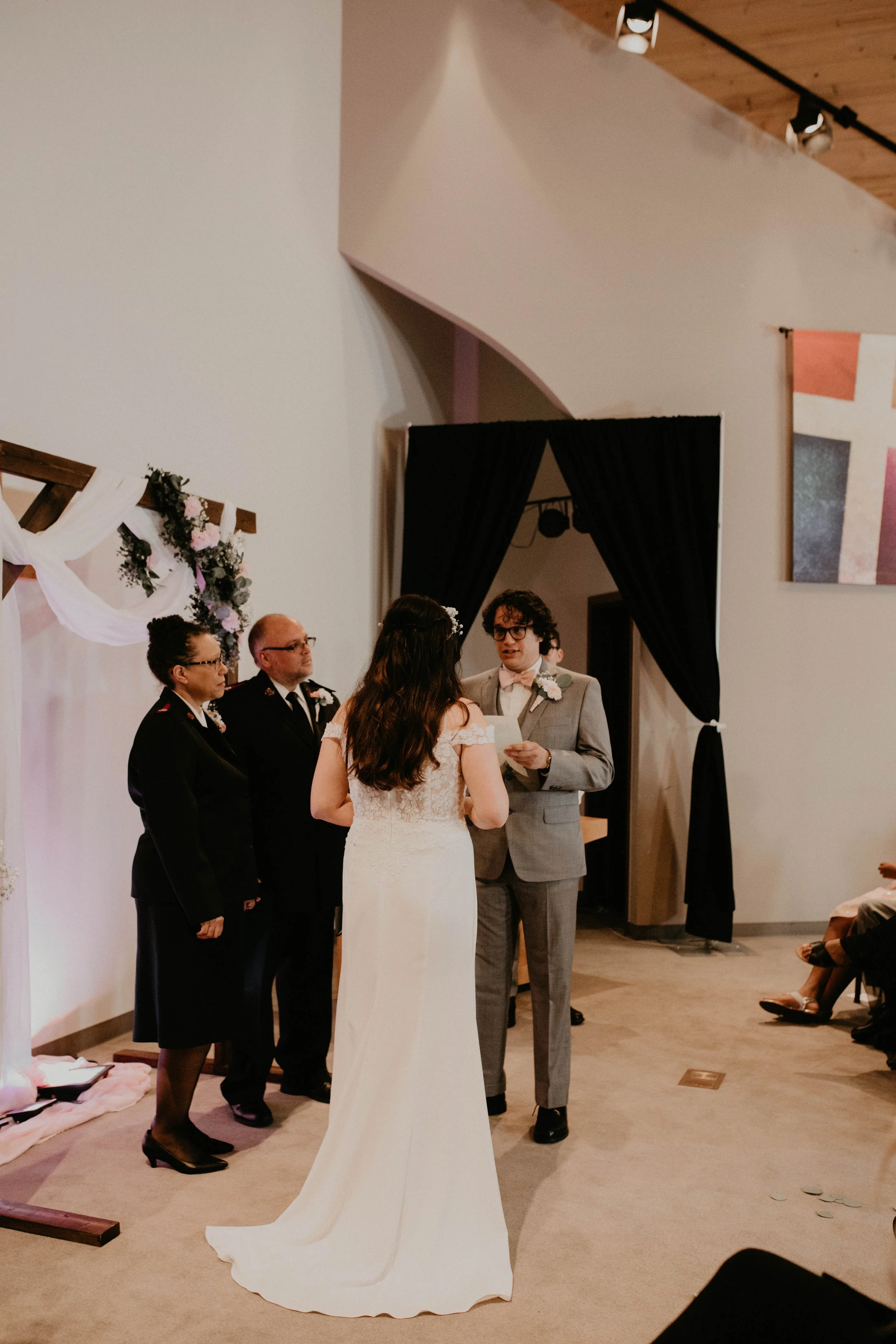 A wedding ceremony with a bride and groom exchanging vows, with two officiants, in a modern indoor venue decorated with flowers and drapery. Seattle, WA wedding photography.