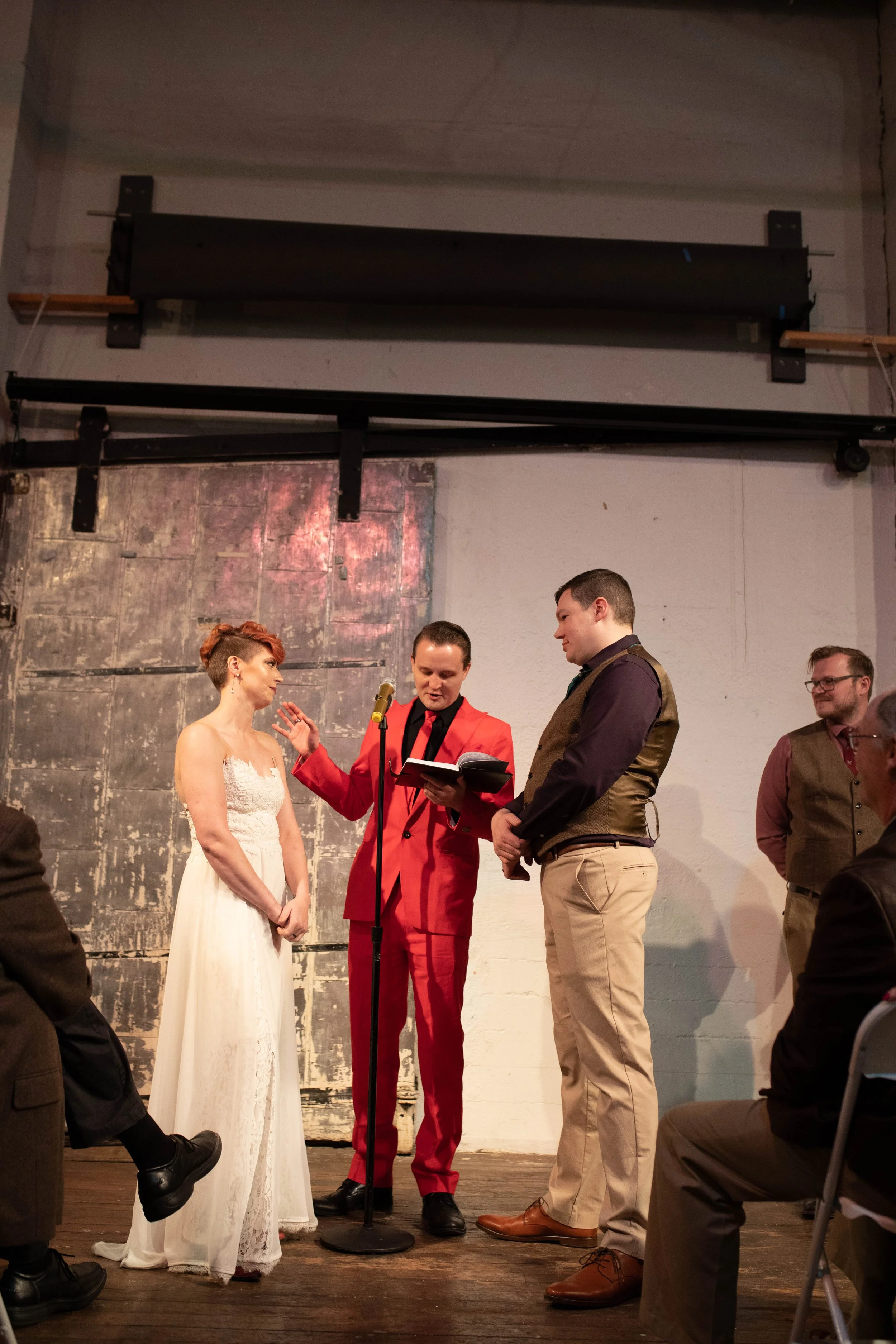 A wedding ceremony with a bride in a white dress and a groom in khakis and a vest standing before a person reading vows or making an announcement, with other guests seated nearby. Pioneer Square, Seattle, WA wedding photography.
