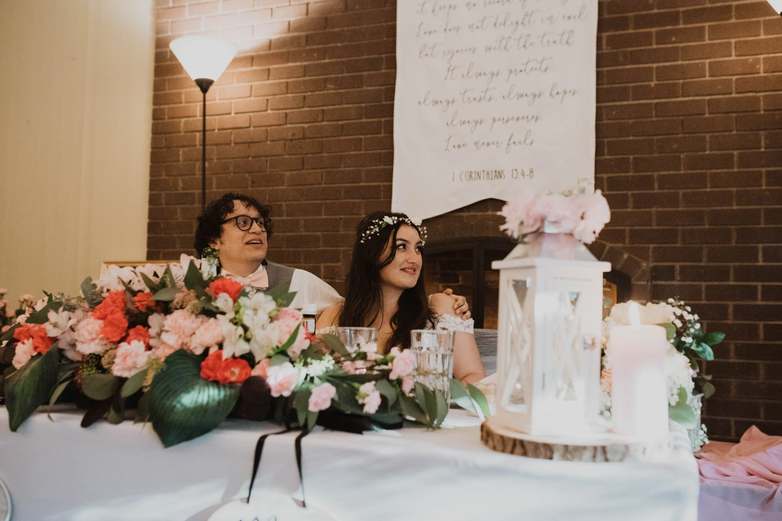 A bride and groom sitting at a wedding reception table decorated with pink and white flowers, a white lantern with pink flowers, glasses of water, and candles, with a brick wall and a white cloth with writing hanging behind them.