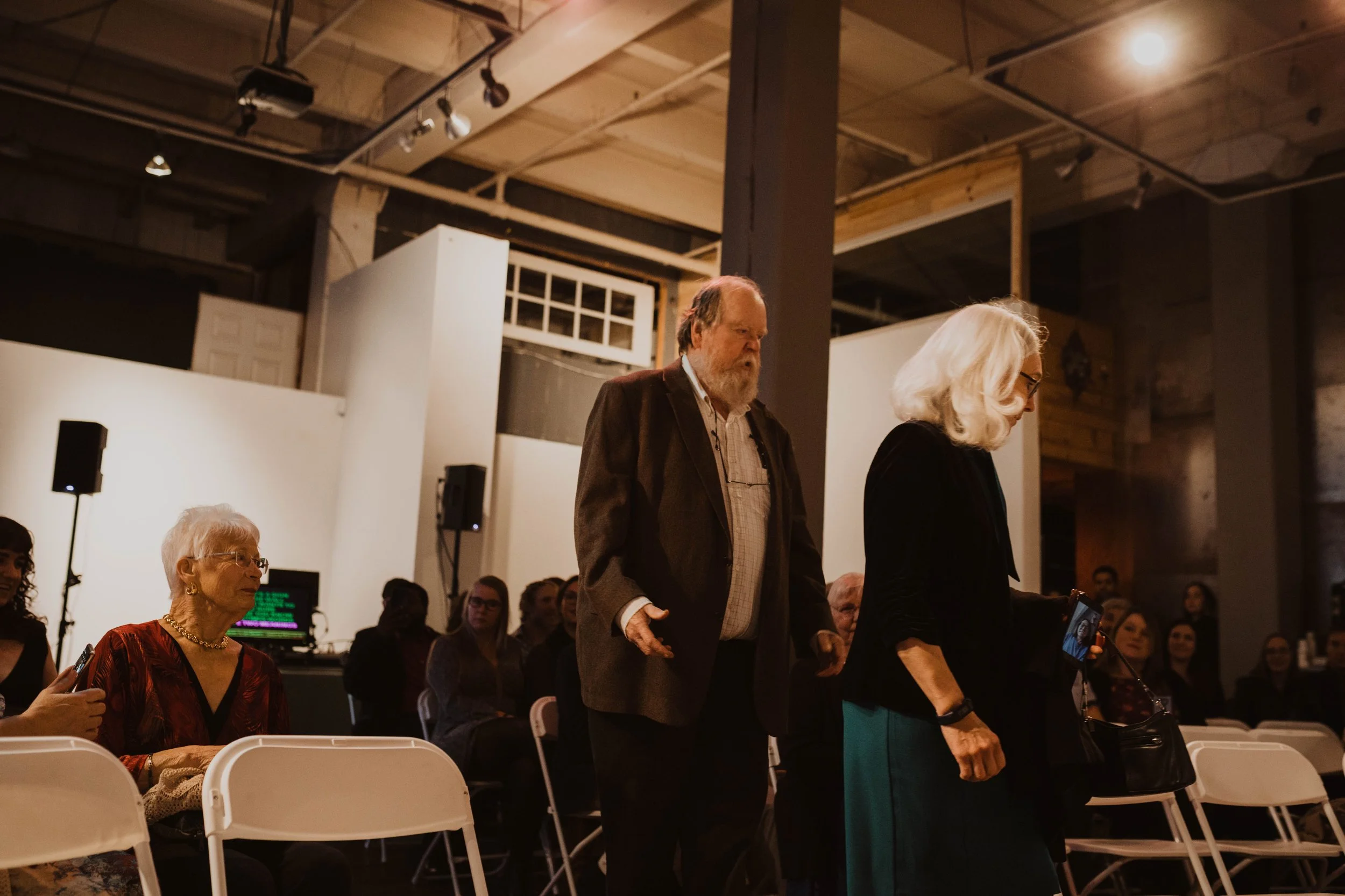 Older woman with white hair wearing glasses, black top, and teal skirt walking down the aisle during an indoor event, with other attendees seated or standing nearby. Pioneer Square, Seattle, WA wedding photography.