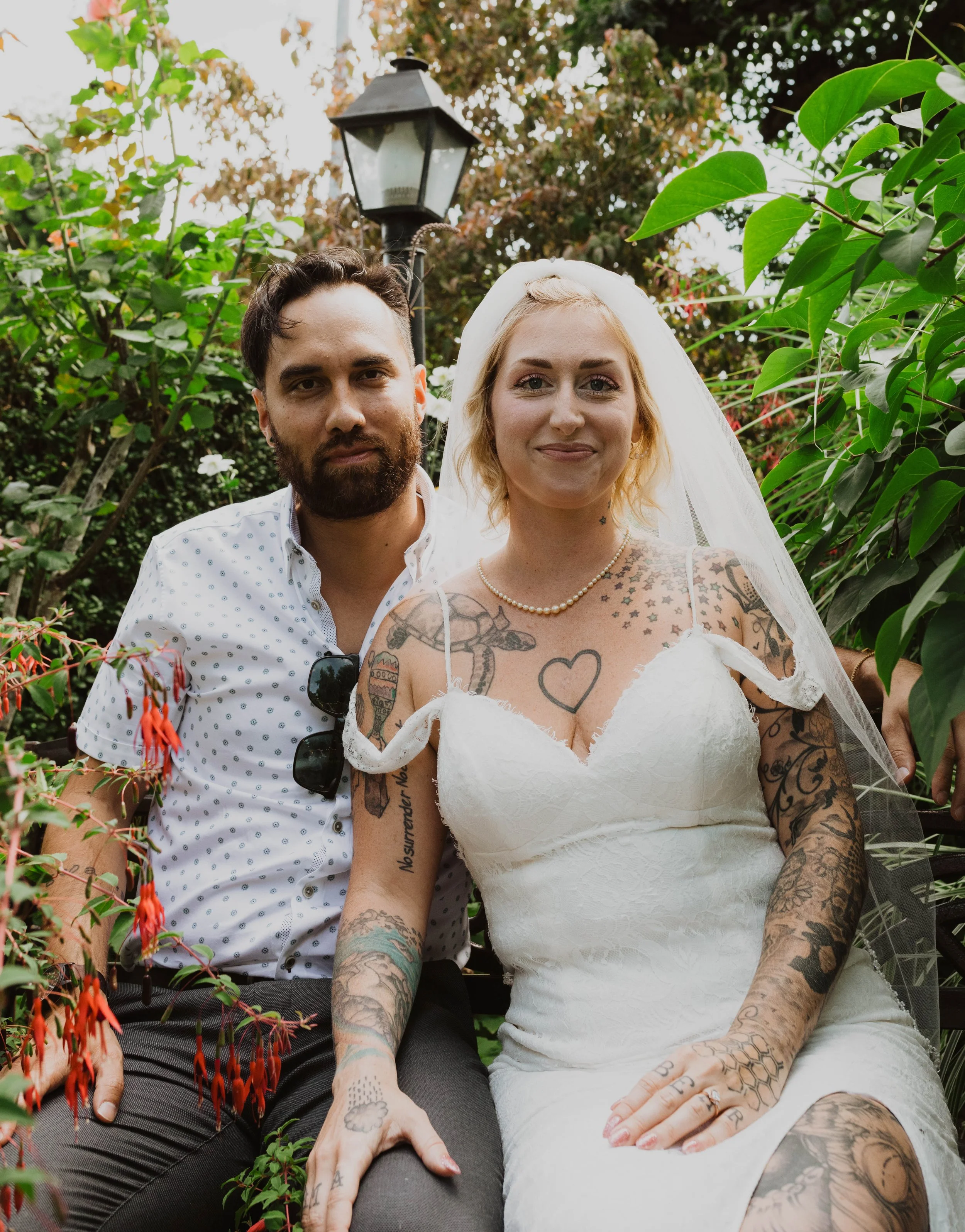 A newlywed couple sitting on a garden bench surrounded by greenery and flowers, with a lamppost in the background. The bride has blonde hair, multiple tattoos, and is wearing a white wedding dress with a veil, while the groom has dark hair and a bear