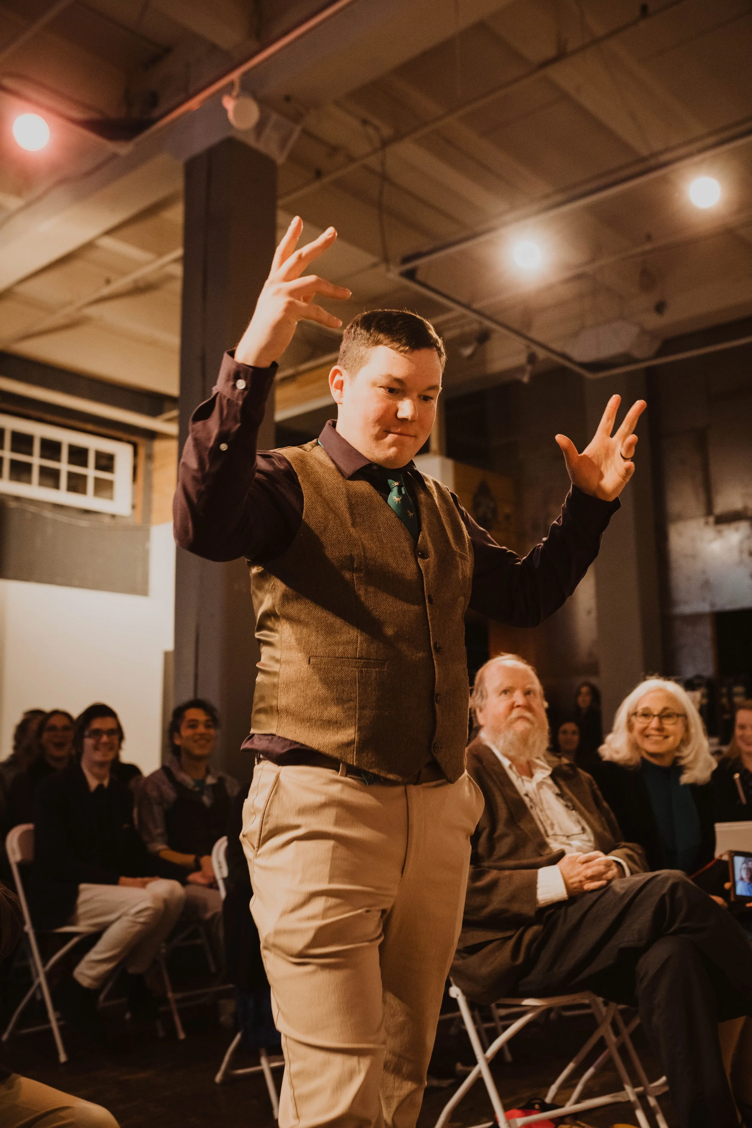 A young man with short dark hair raising his hands in a gesture during a presentation or performance, with an audience seated behind him in a dimly lit, industrial-style room. Pioneer Square, Seattle, WA wedding photography.
