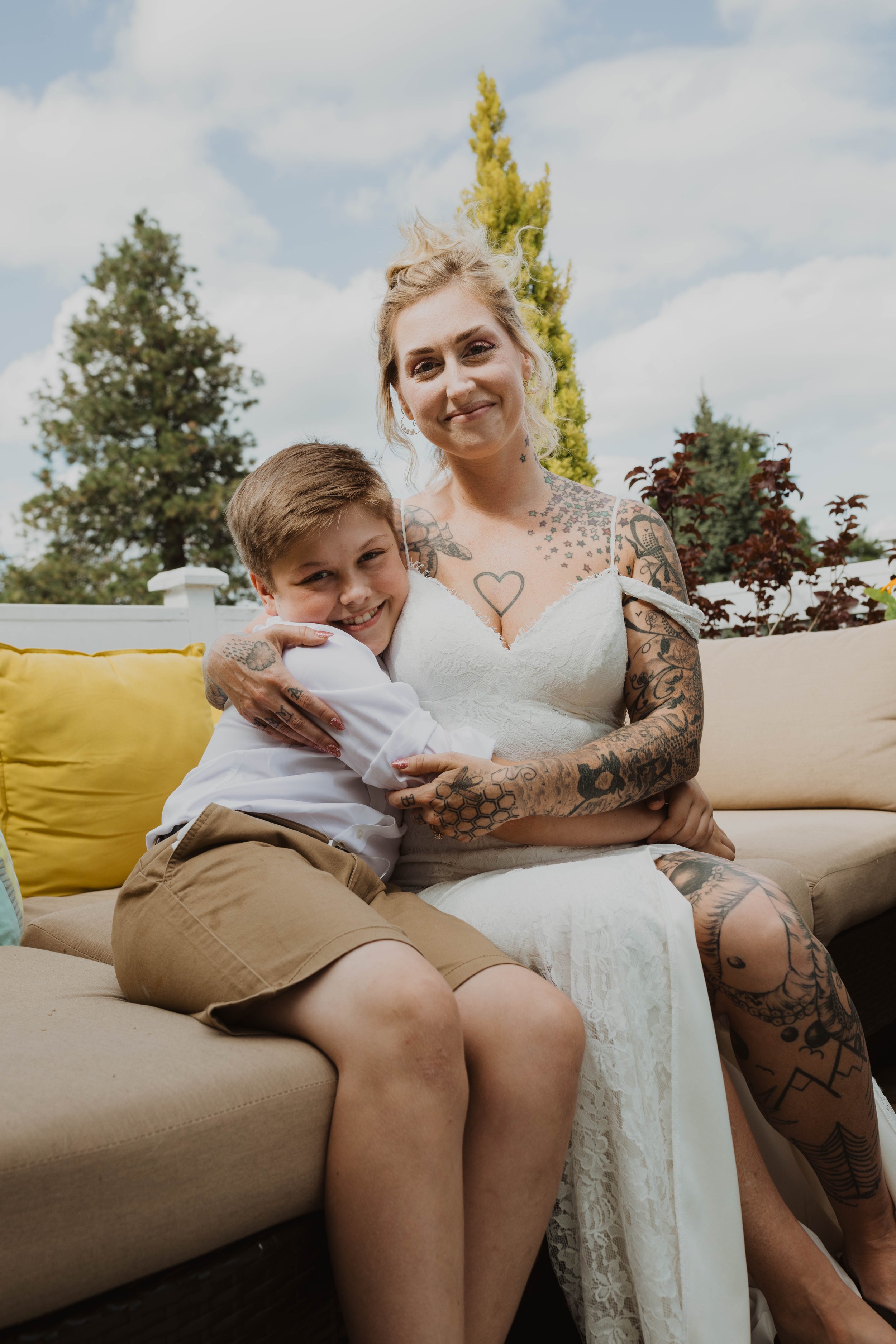 A woman with tattoos and a boy sitting on her lap enjoying an outdoor moment, with trees and cloudy sky in the background. Seattle, WA wedding photography.