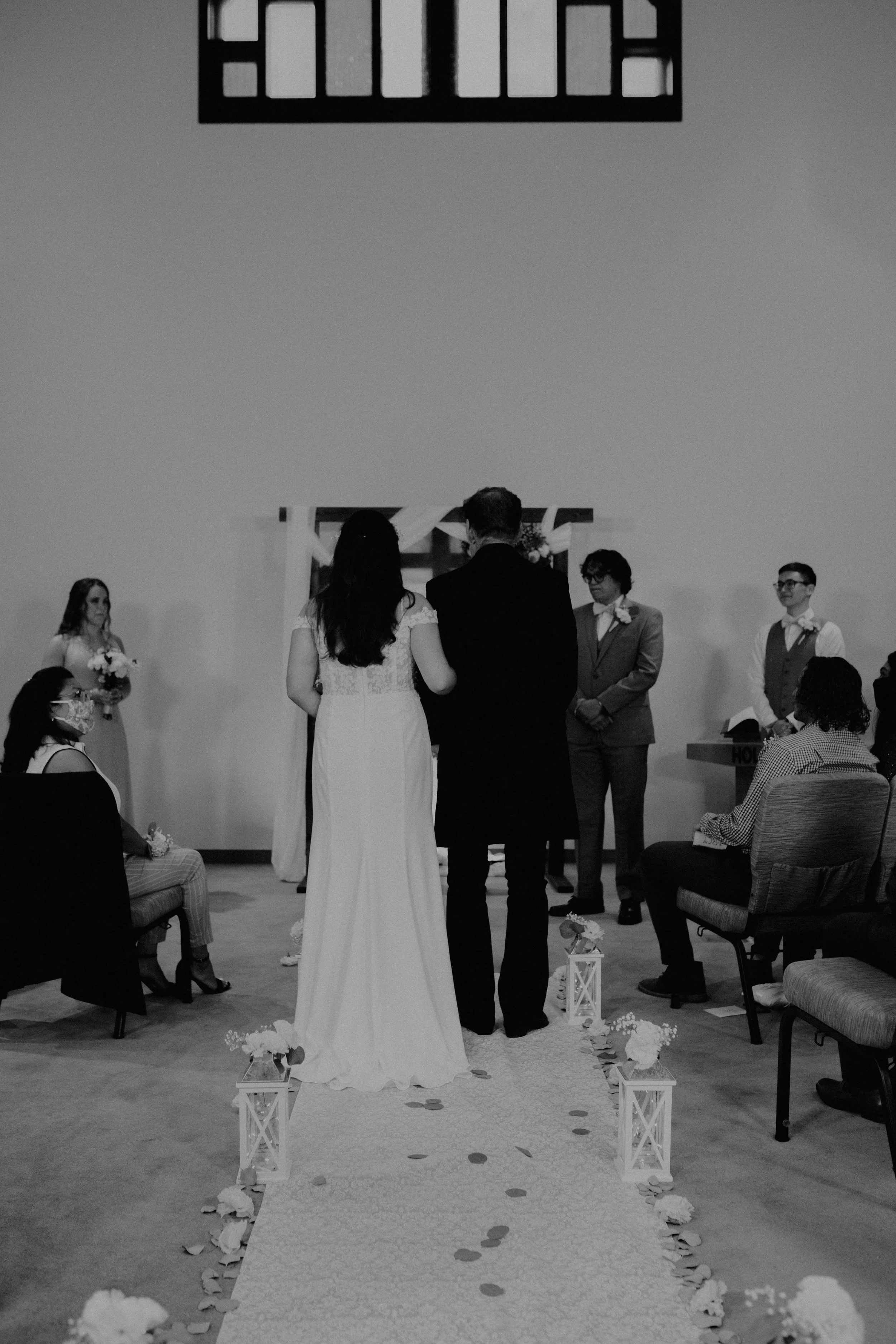 Black and white photo of a wedding ceremony with the bride and groom standing at the altar, facing the officiant, during the vows, with seated and standing guests watching. Seattle, WA wedding photography.