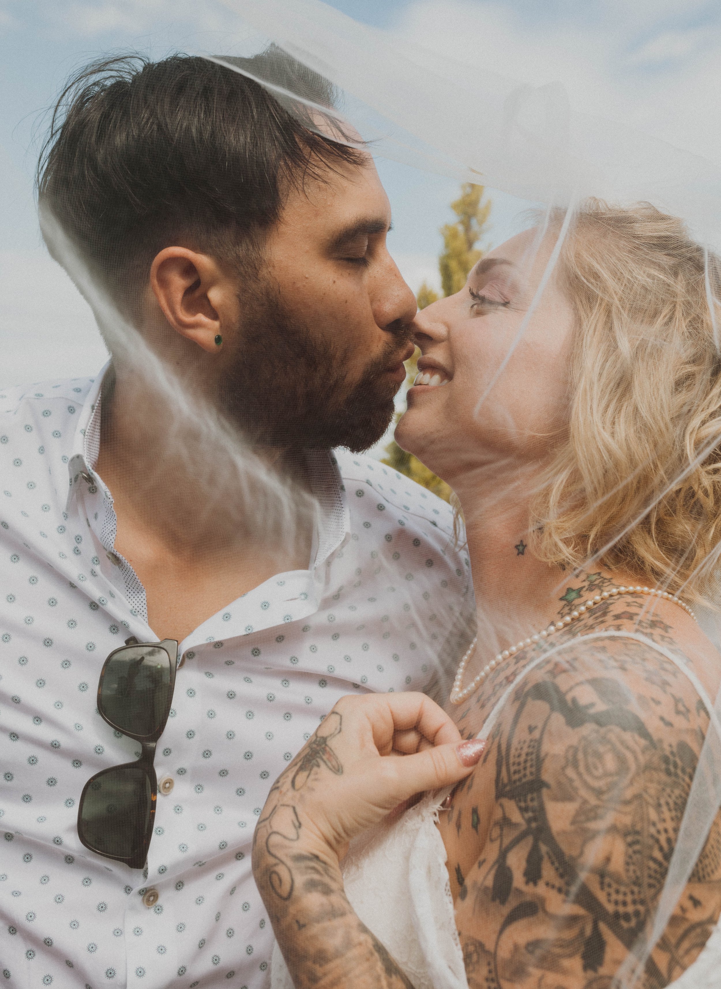 A couple showing a close-up moment, about to kiss, standing outdoors with a blue sky and trees in the background. The woman has tattoos, blonde hair, and is wearing a pearl necklace, while the man has a beard, is wearing a white shirt with small patt