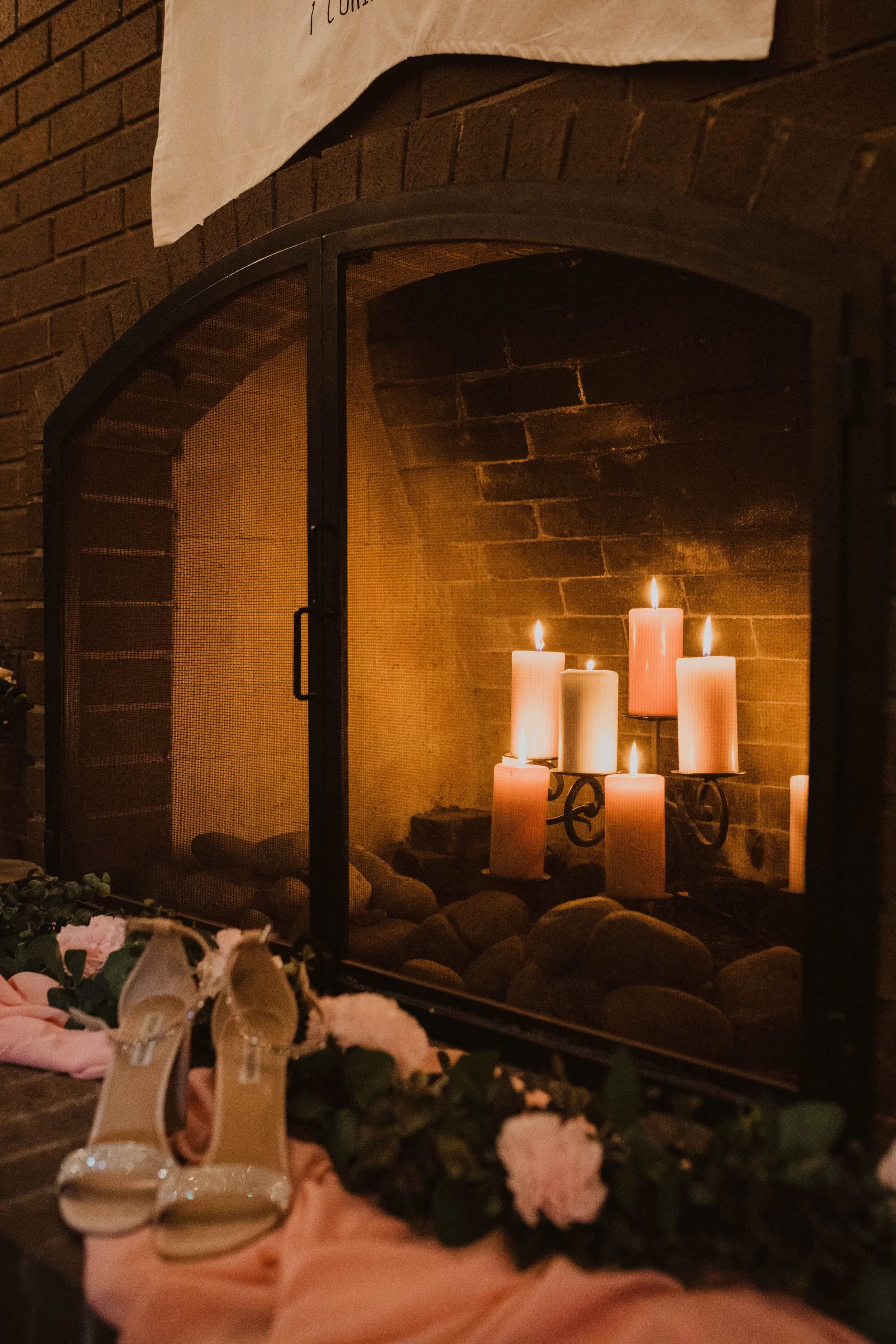 Candles glowing inside a brick fireplace, with pink and white flowers and beige high-heeled shoes with ankle straps in front. Seattle, WA wedding photography.