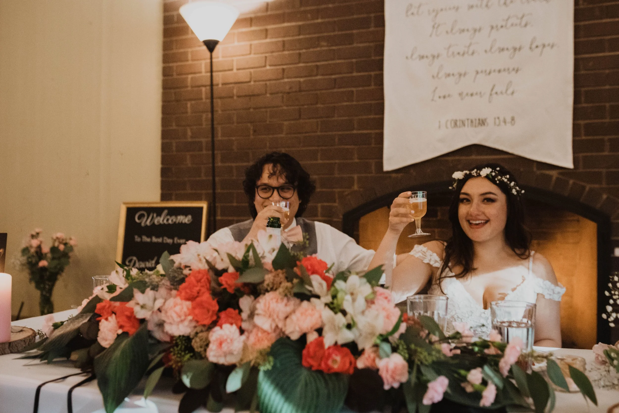 A bride and groom sitting at a wedding reception table, raising drinks and smiling. The table is decorated with a large floral centerpiece of pink and white flowers, and there is a sign that says 'Welcome to the Best Day Ever' on the table. The backg