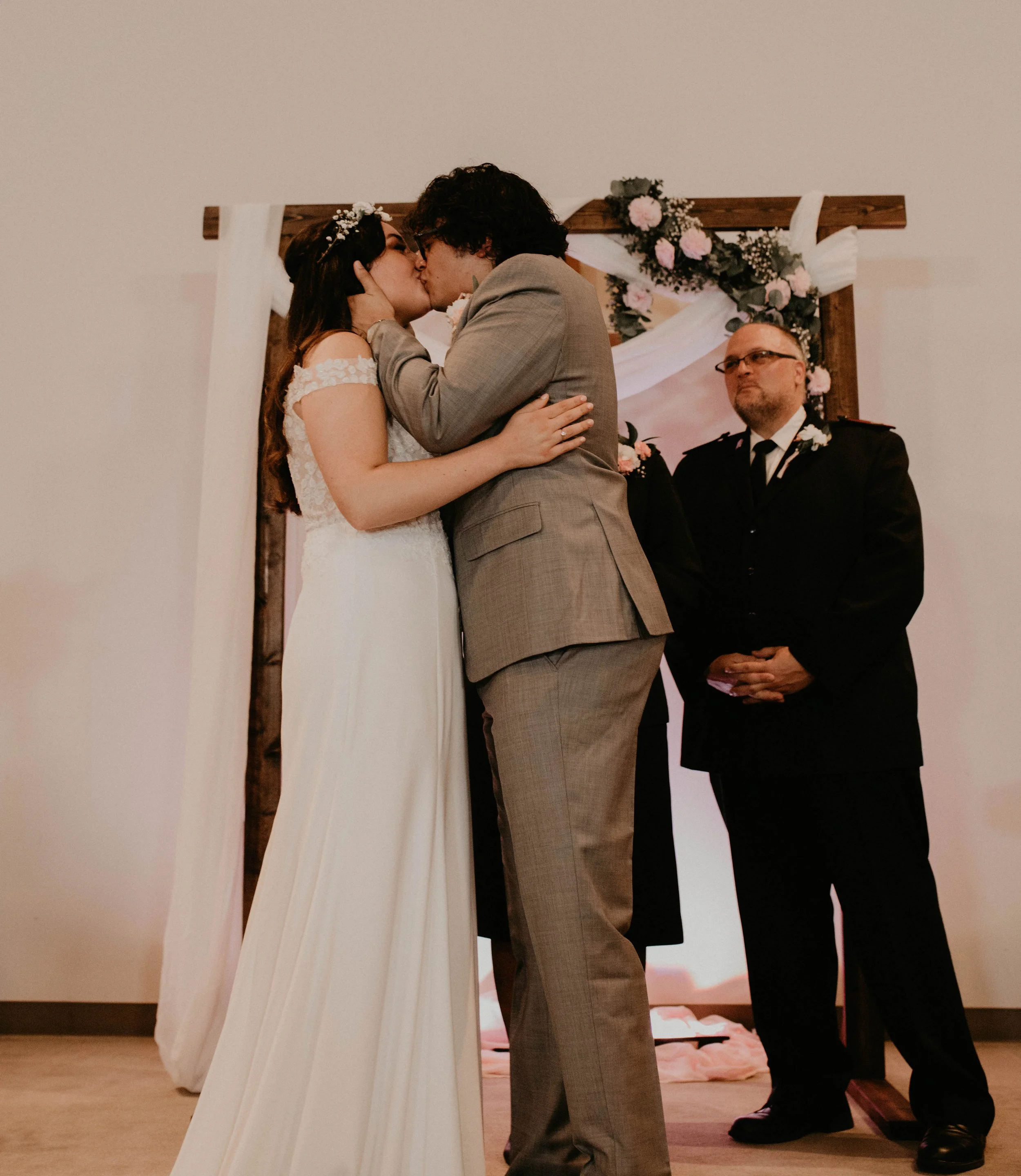 A couple sharing a kiss at their wedding ceremony with an officiant standing nearby, wedding arch with pink and white flowers in the background. Seattle, WA wedding photography.