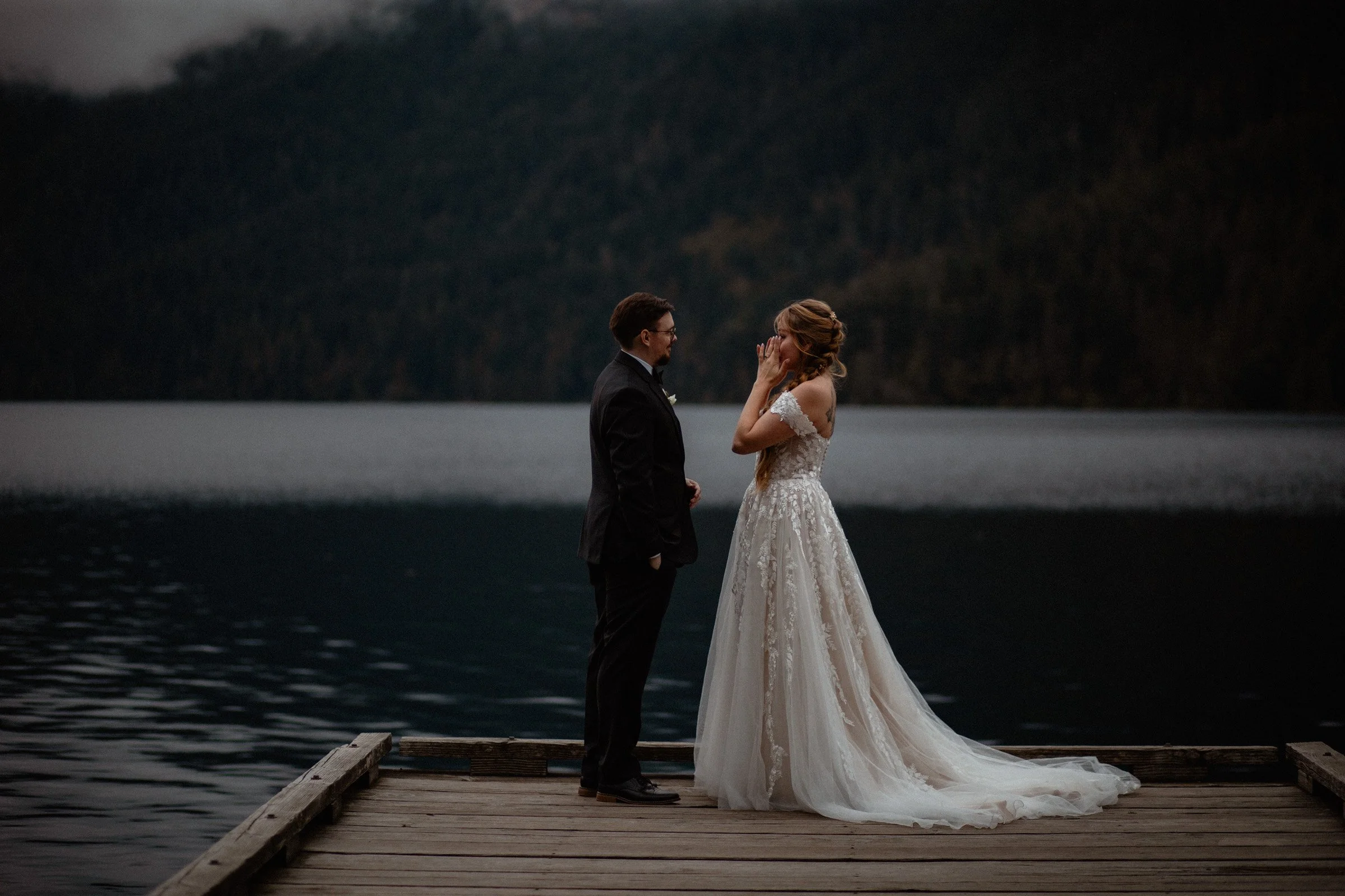Bride and groom standing on a dock at Lake Crescent with mountains reflected in the water during their Lake Crescent Lodge wedding in Port Angeles, WA.
