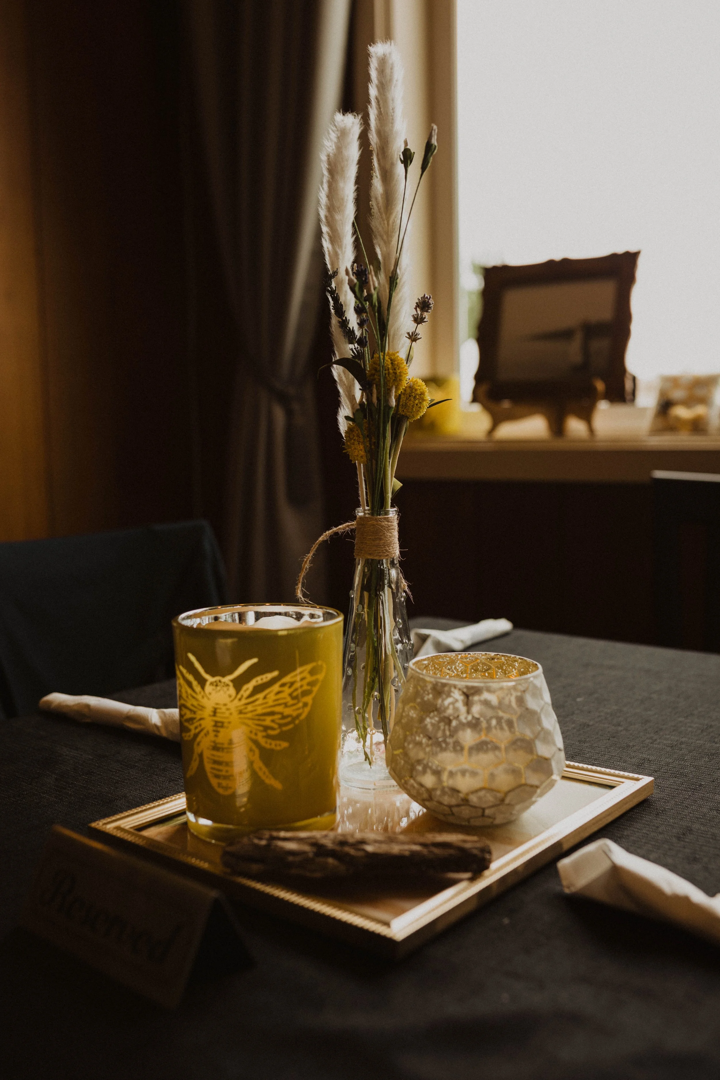 Table centerpiece with a glass jar of dried flowers, a yellow candle with a bee illustration, and a honeycomb textured candle holder on a wooden tray, set on a dark table with napkins. Long Beach, WA wedding photography.