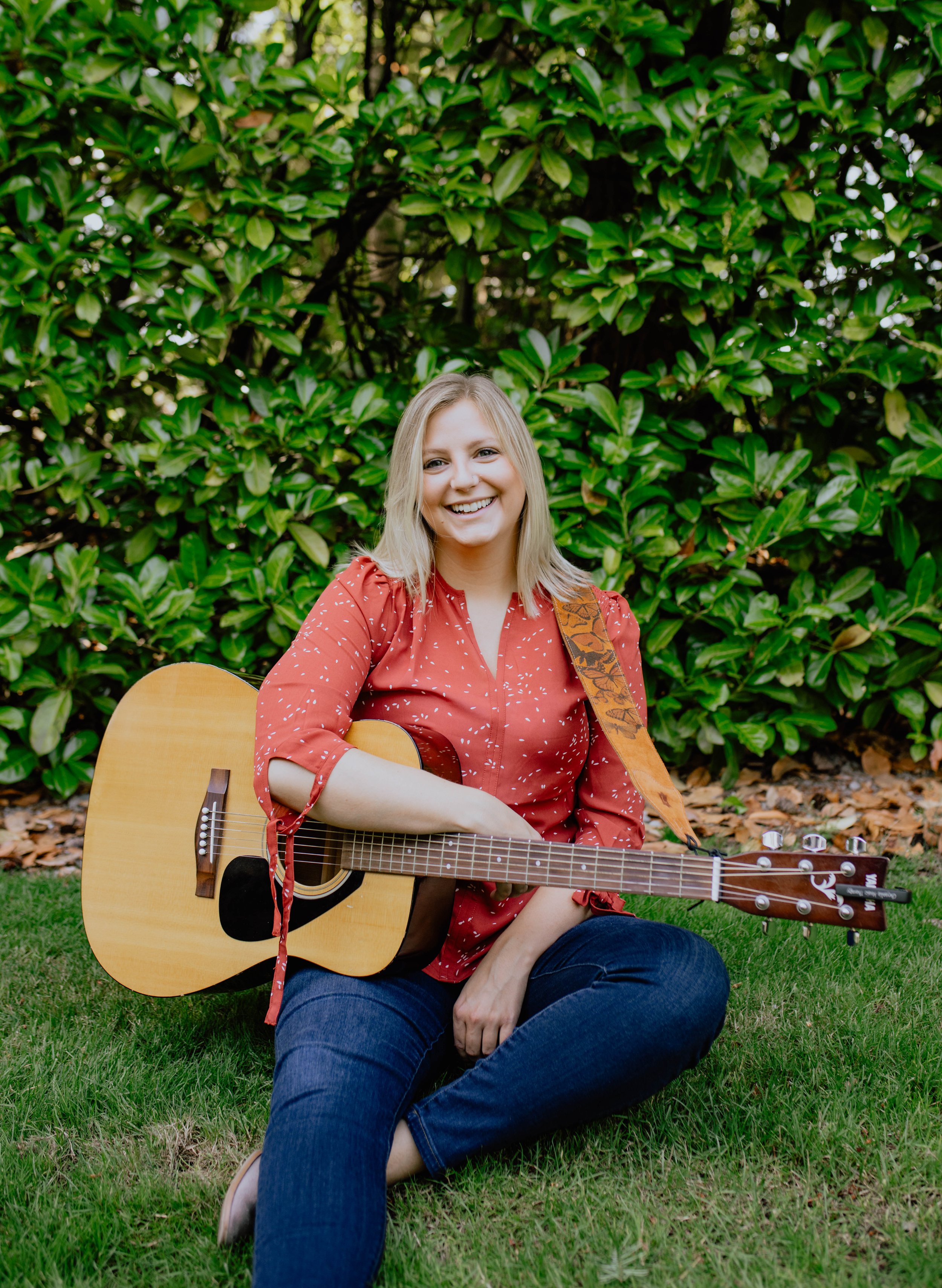 A woman with blonde hair, wearing a red blouse and jeans, sitting on the grass with a guitar, smiling at the camera, with green bushes in the background. Seattle professional head shot photography