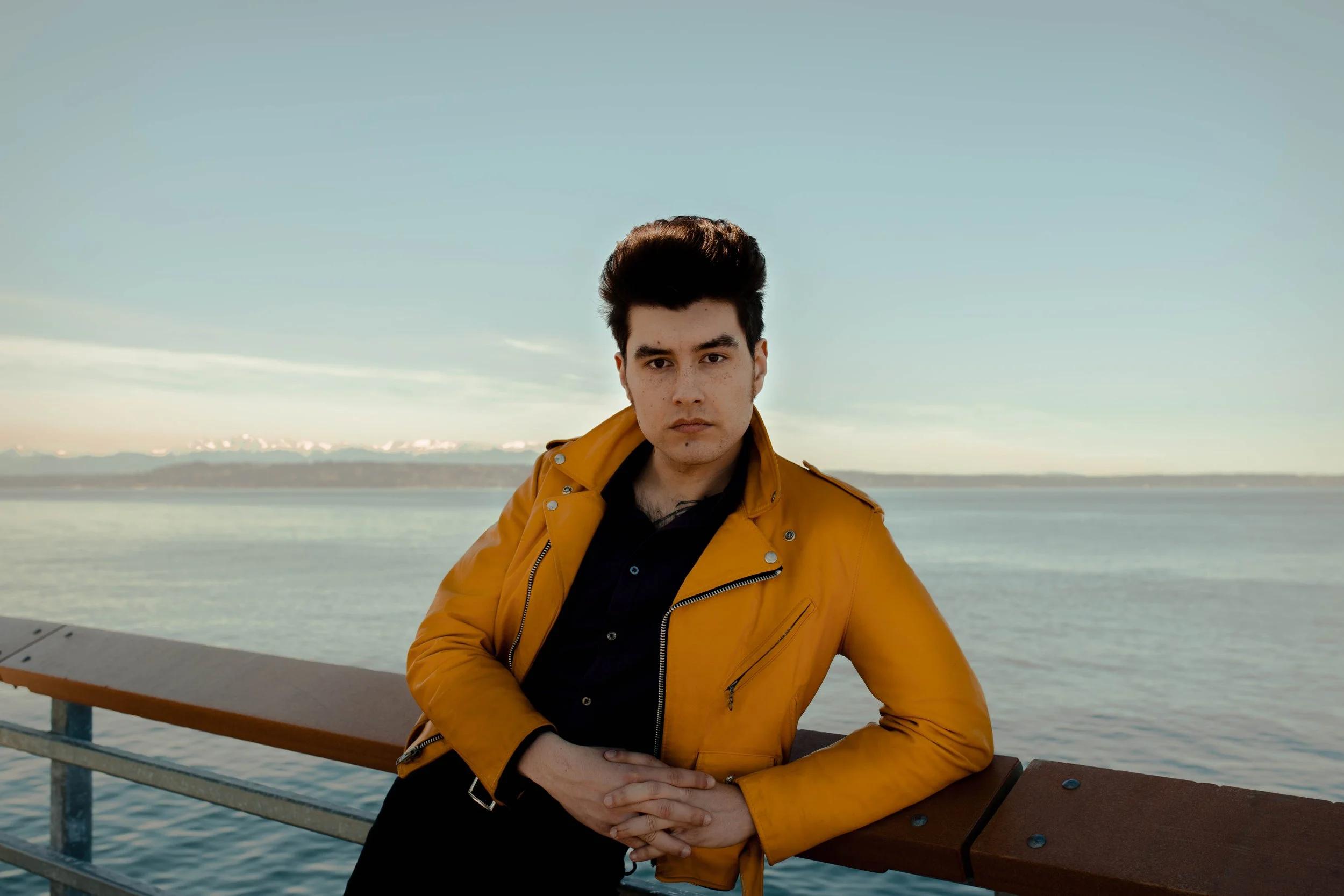 A young man with dark hair and pale skin wearing a yellow leather jacket and black shirt leans on a wooden railing by a body of water, with a distant shoreline and snow-capped mountains in the background. Seattle professional head shot photography