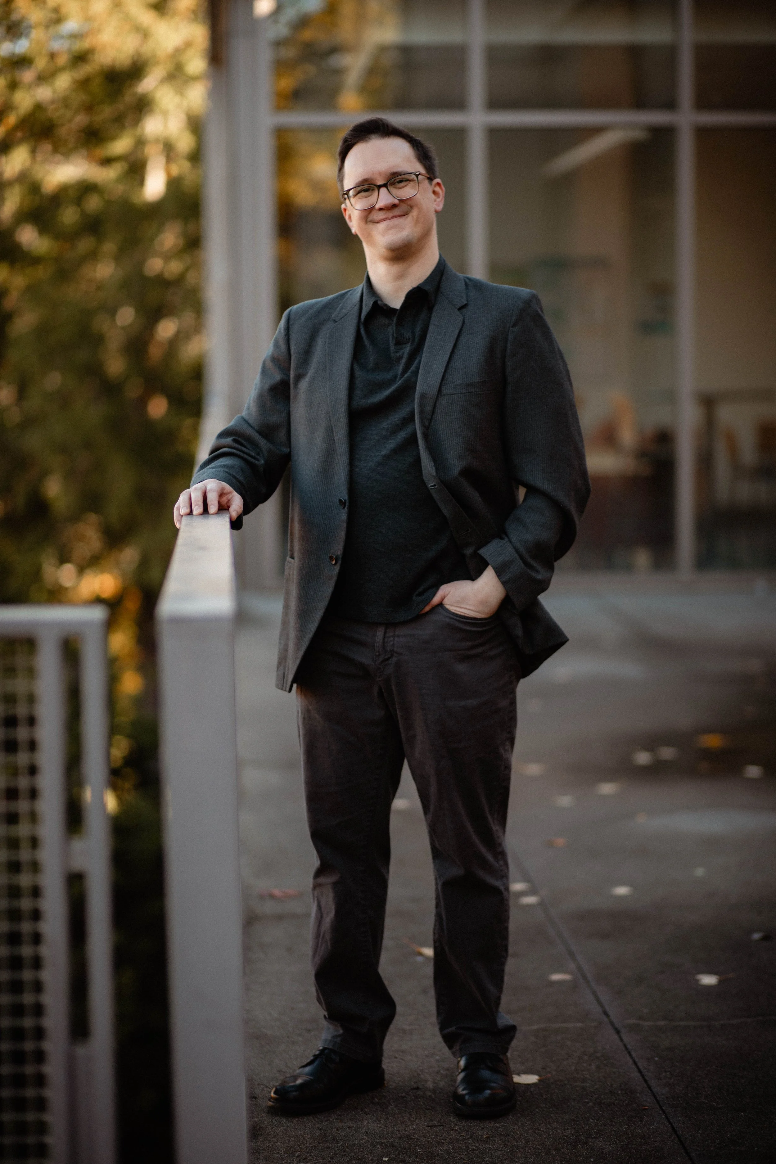 A man with glasses, wearing a dark blazer and black shirt, leaning on a railing outdoors with trees and a building in the background. Seattle professional head shot photography
