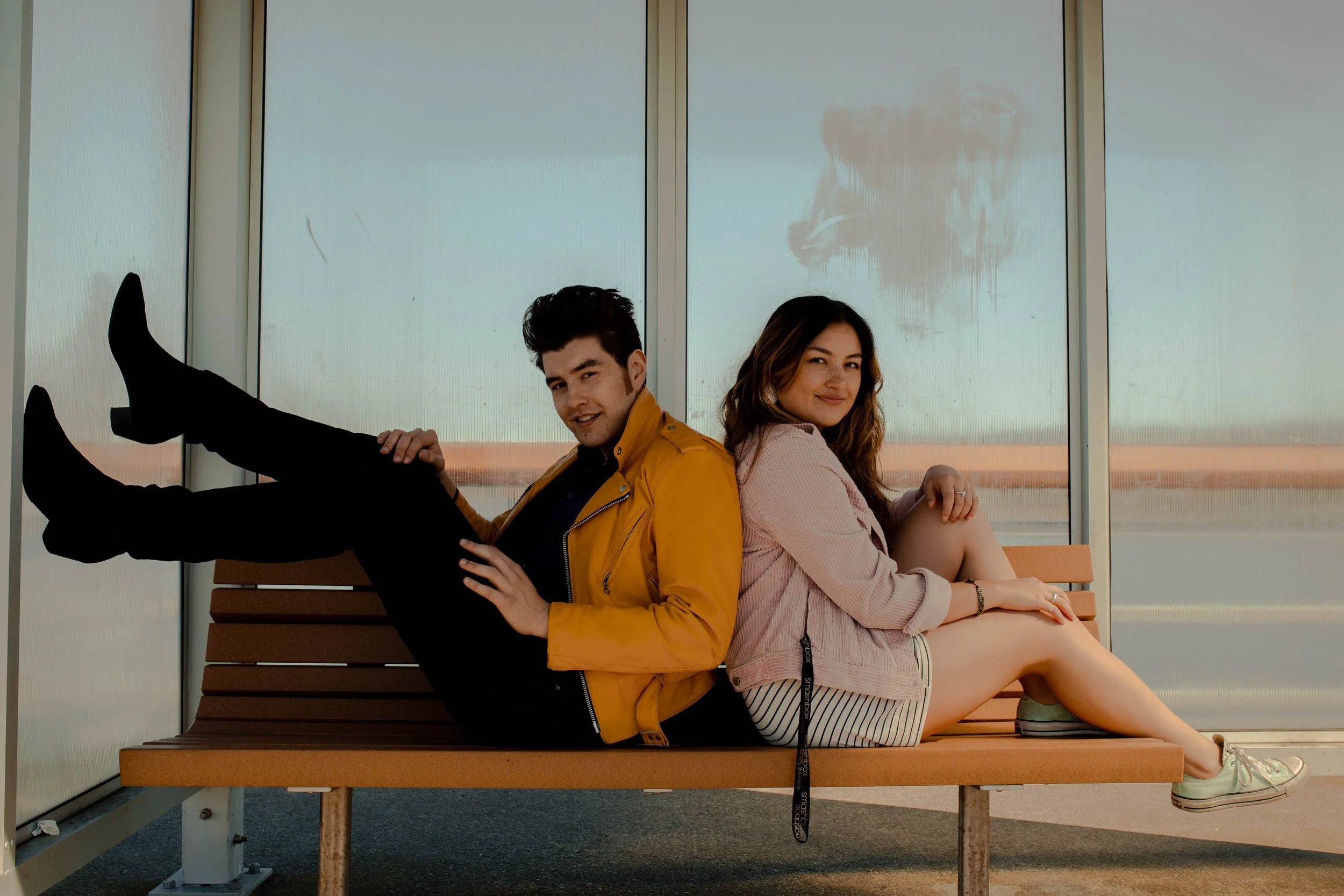 A man and a woman sitting back-to-back on a bench at an airport. The man is leaning slightly back with his legs extended and crossed at the ankles, wearing a yellow jacket and black pants. The woman sits with her legs extended forward, wearing a pink