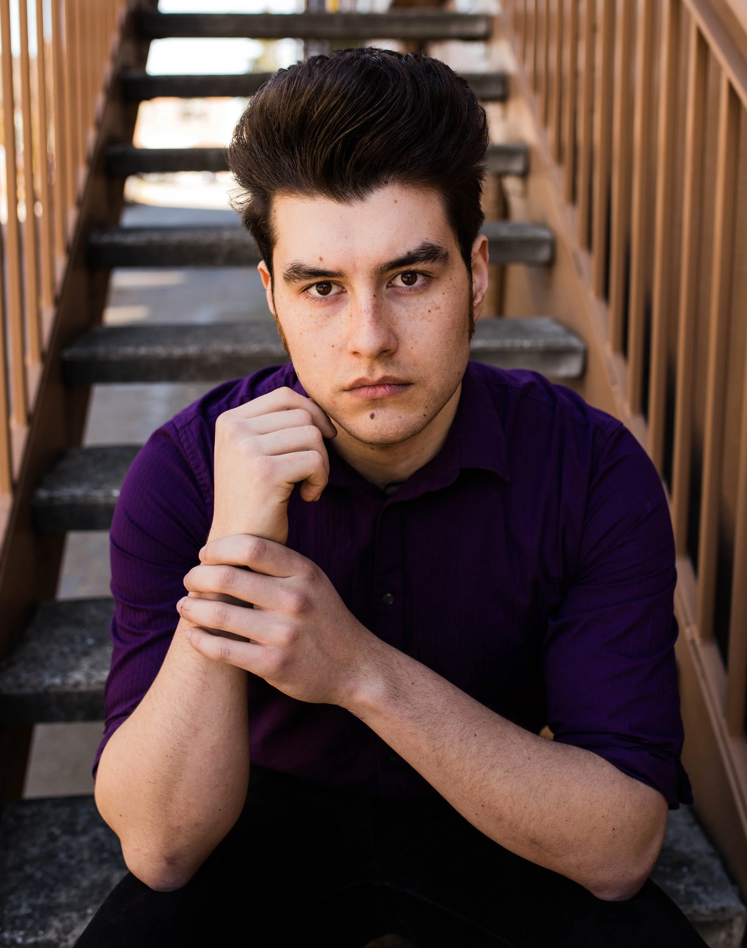 A young man with dark hair and freckles sits on outdoor stairs, resting his chin on his hand, wearing a purple shirt with a serious expression. Seattle professional head shot photography