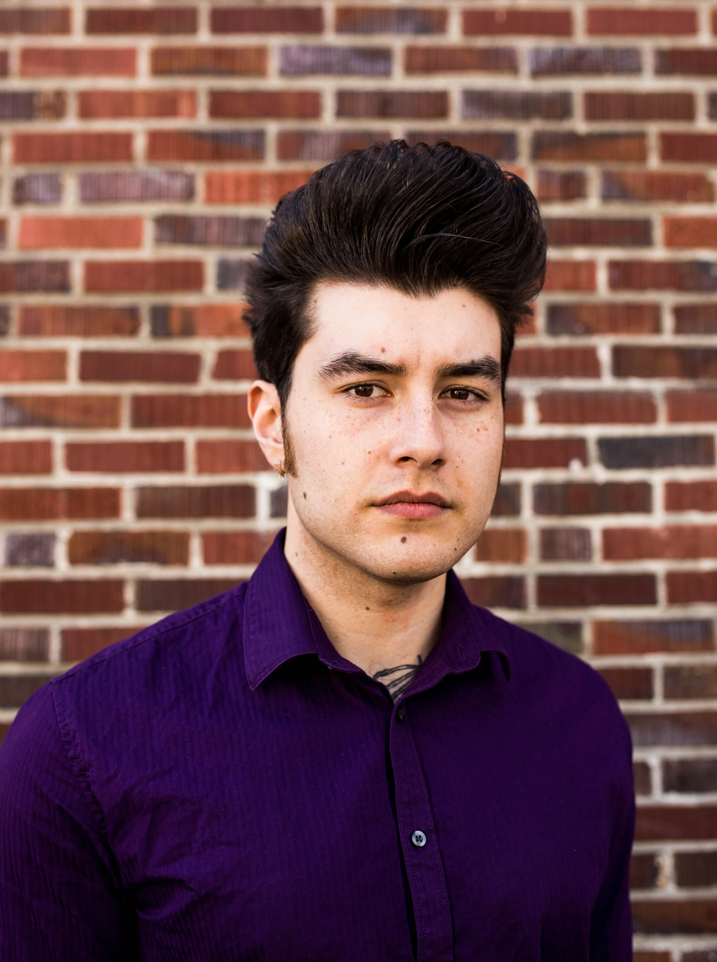 A young man with dark hair and freckles, wearing a purple shirt, standing in front of a brick wall. Seattle professional head shot photography