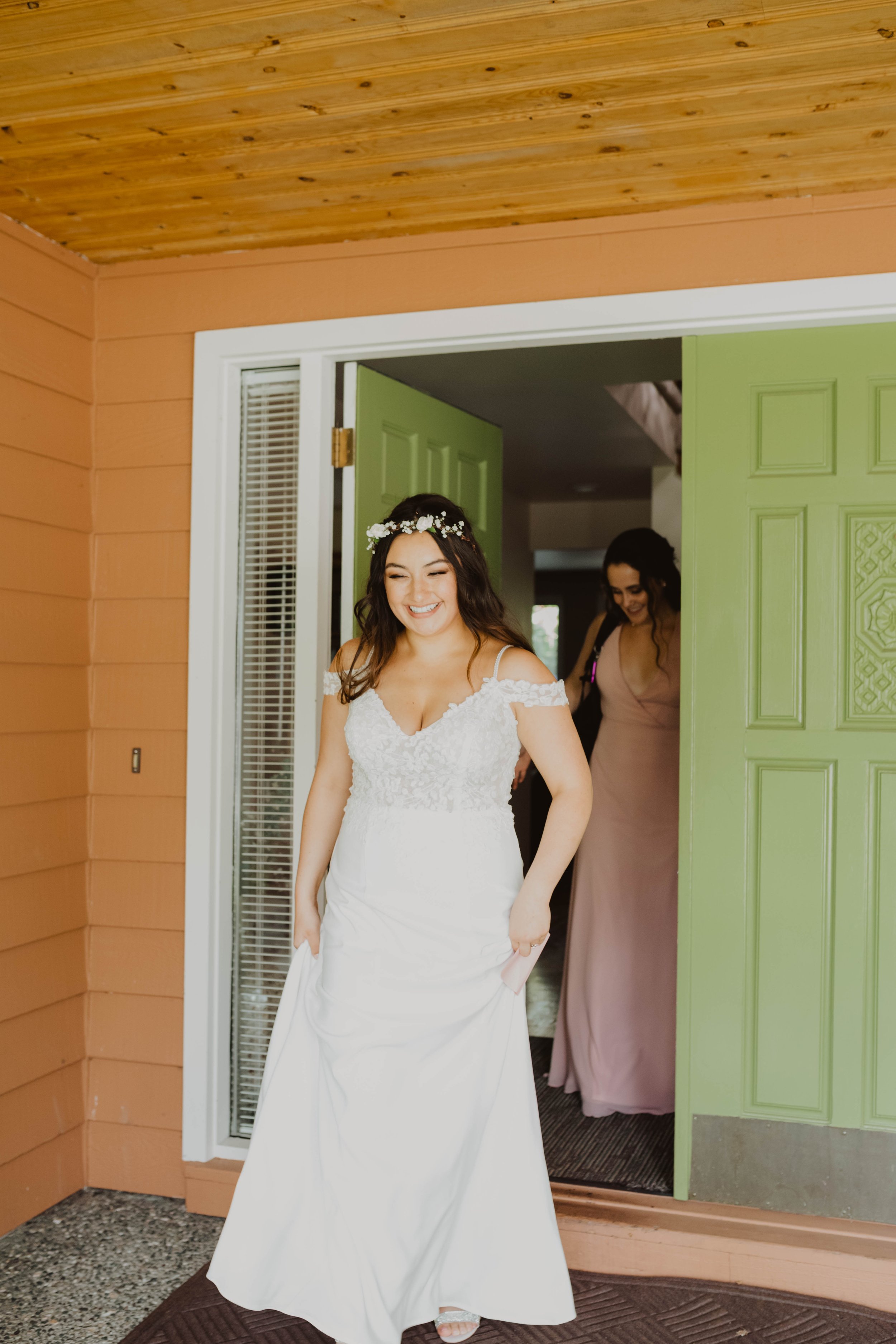 A smiling woman in a white wedding dress, holding her dress, standing at the doorway of a house with a green door, inside a room with wooden ceiling and orange wall paneling, with another woman in a pink dress behind her. Seattle, WA wedding photogra