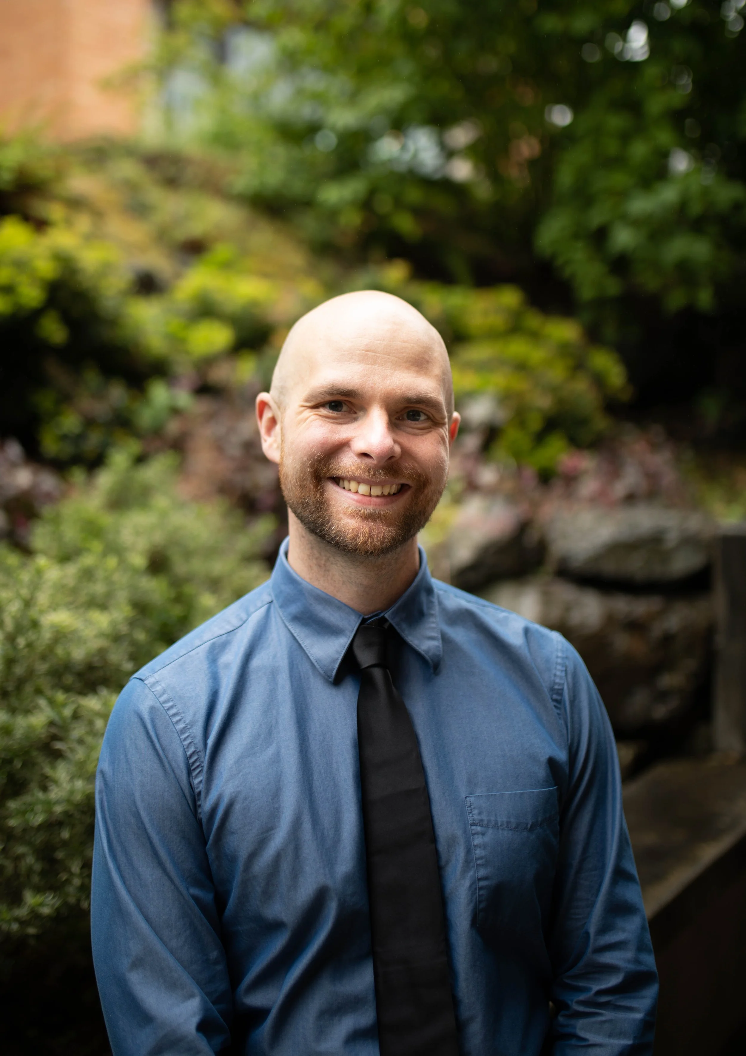 Smiling man with a beard in a blue shirt and black tie outdoors with greenery and rocks in the background. Seattle professional head shot photography
