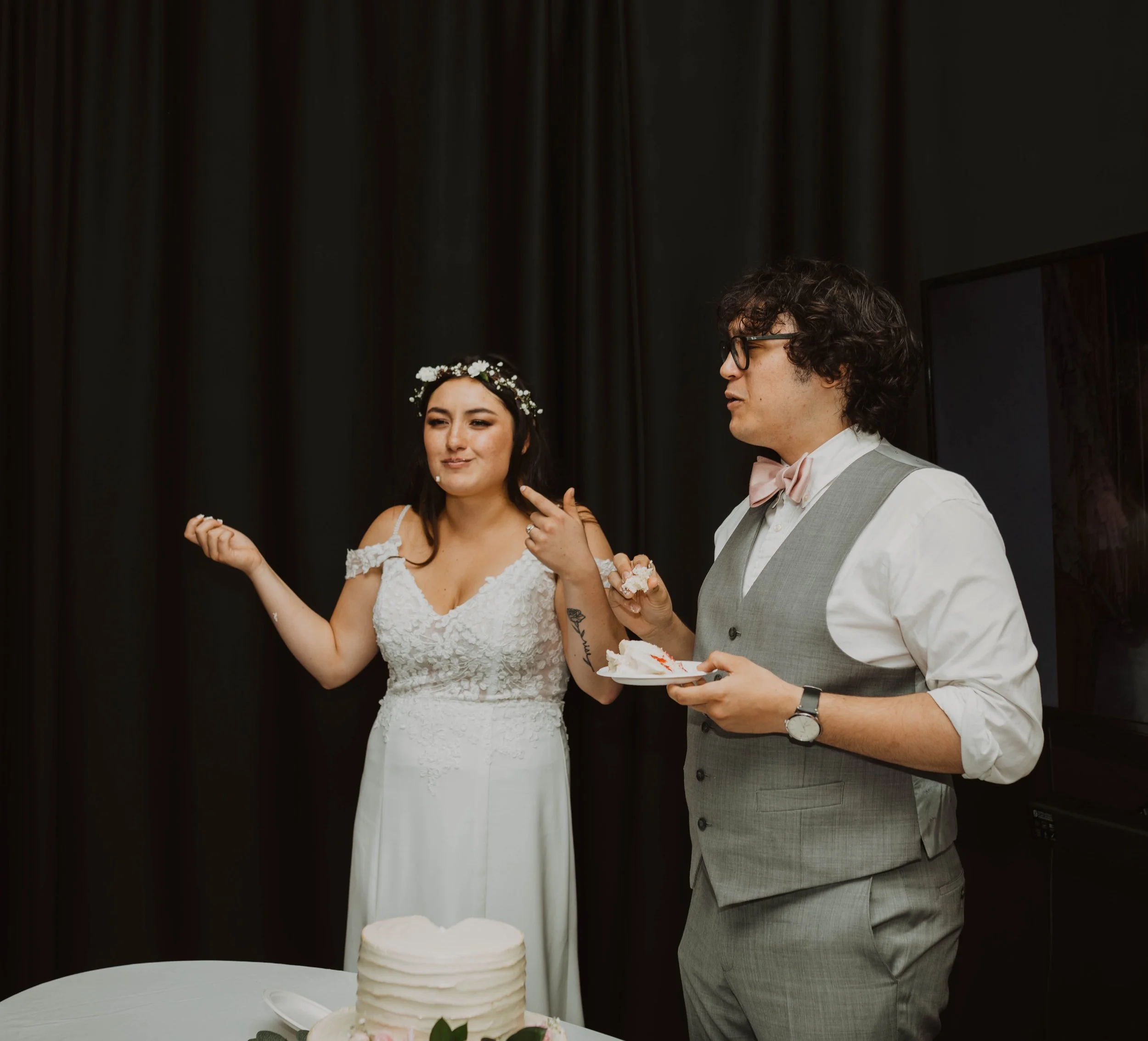 A bride and groom celebrating a wedding with a cake and dessert, the bride wearing a white wedding dress with floral headpiece, the groom in a light grey suit, holding a plate with cake, in front of a black curtain. Seattle, WA wedding photography.