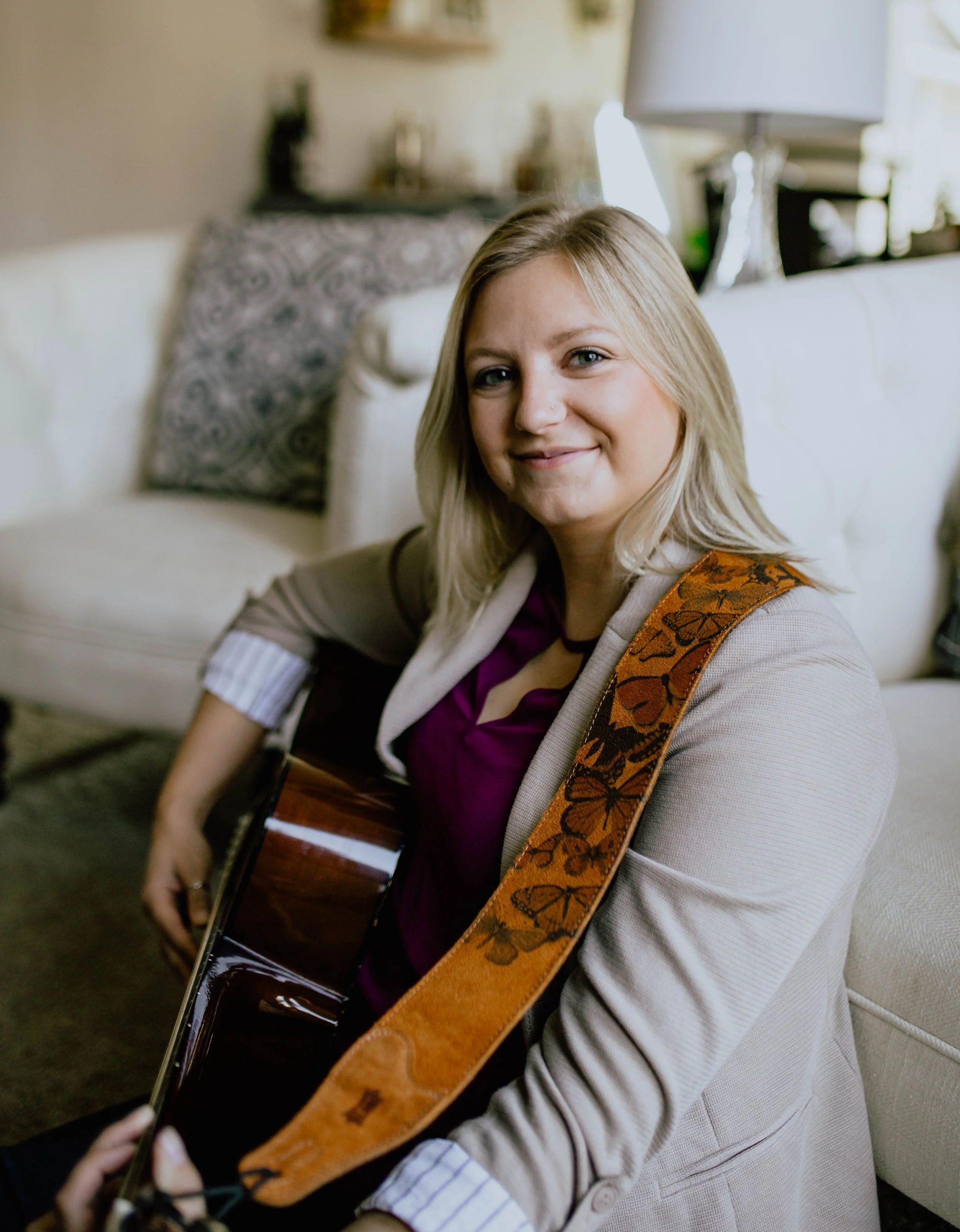 A woman with blonde hair, wearing a beige blazer and purple top, sitting on a sofa with a guitar on her shoulder, in a cozy living room. Seattle professional head shot photography