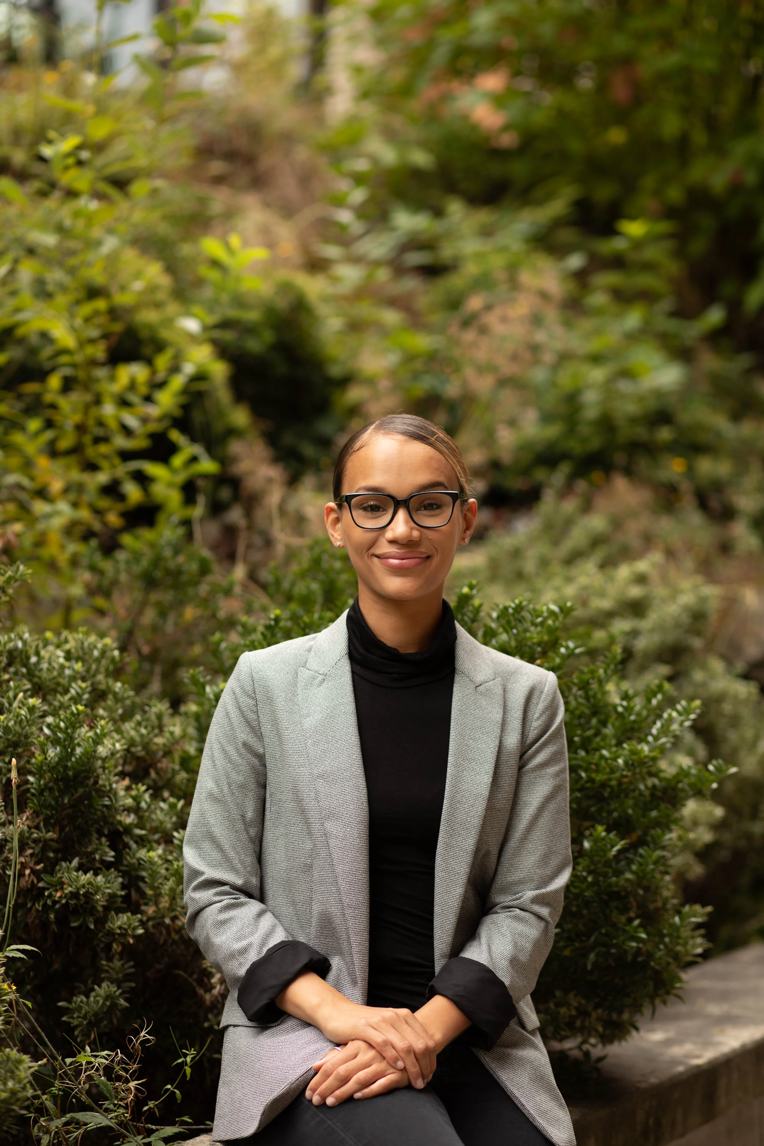 A young woman with glasses and a ponytail sitting outdoors among green bushes, wearing a gray blazer and black turtleneck, smiling at the camera. Seattle professional head shot photography