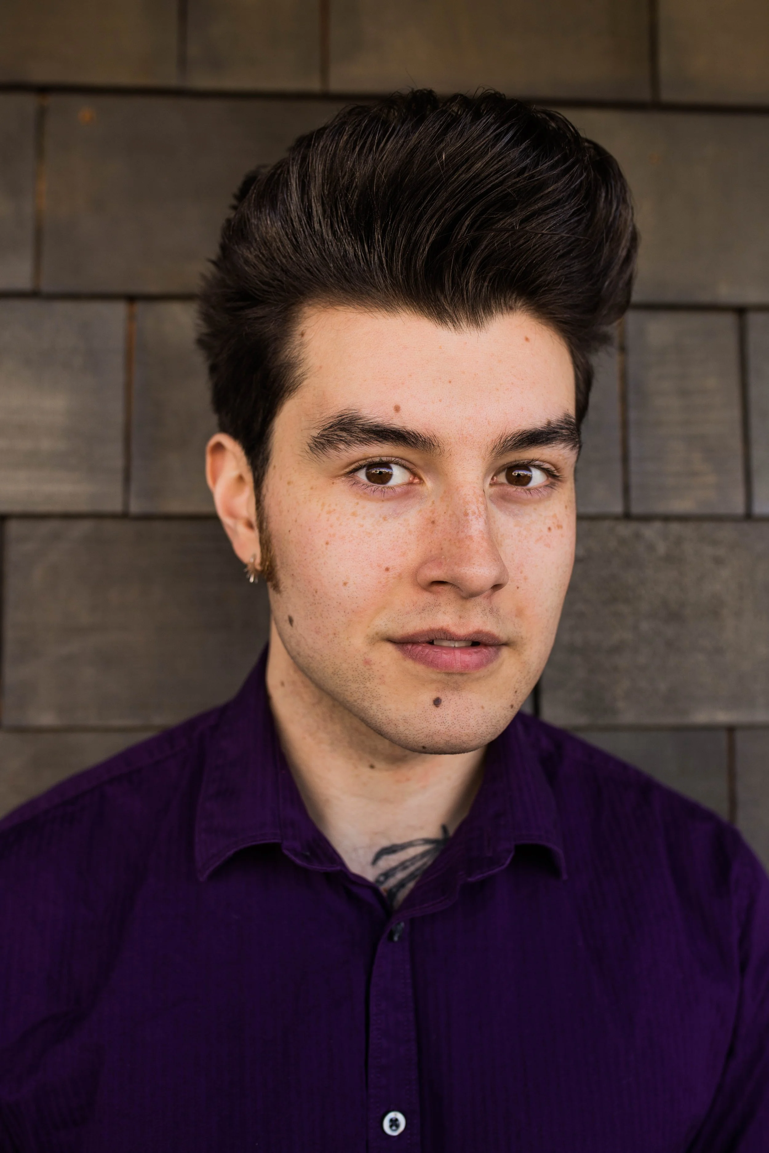 A young man with dark hair styled in a pompadour, light skin with freckles, wearing a dark purple shirt, standing in front of a dark wooden wall. Seattle professional head shot photography