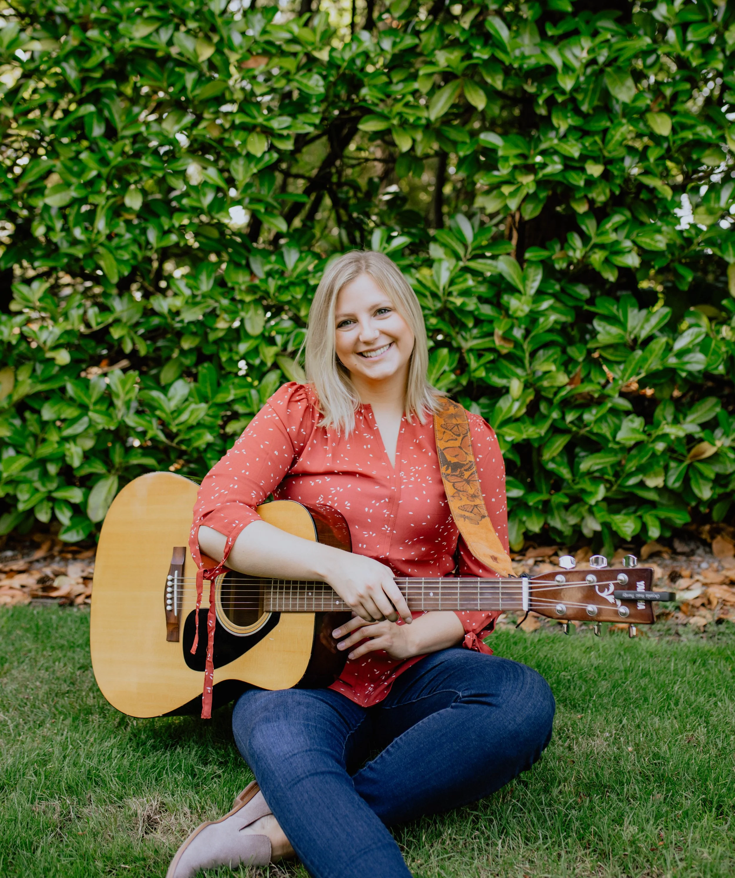 A woman with blonde hair sitting on grass in front of green bushes, smiling, holding an acoustic guitar. Seattle professional head shot photography