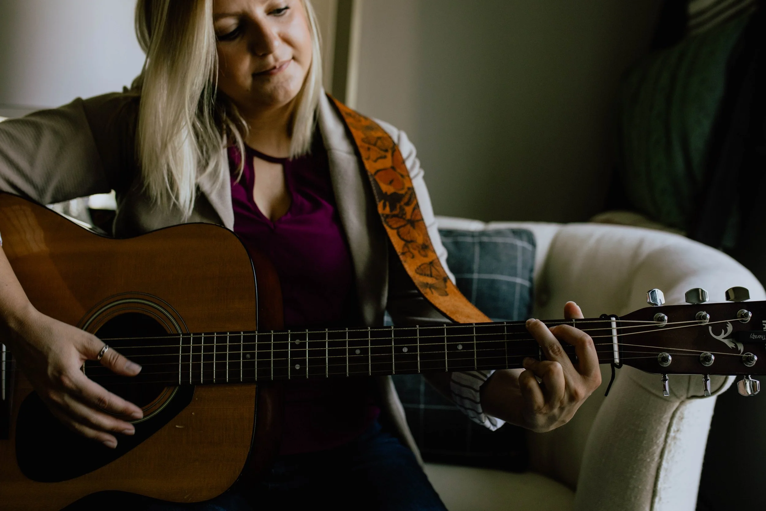 A woman playing an acoustic guitar indoors, sitting on a beige couch with a plaid pillow, with a window partially visible in the background. Seattle professional head shot photography