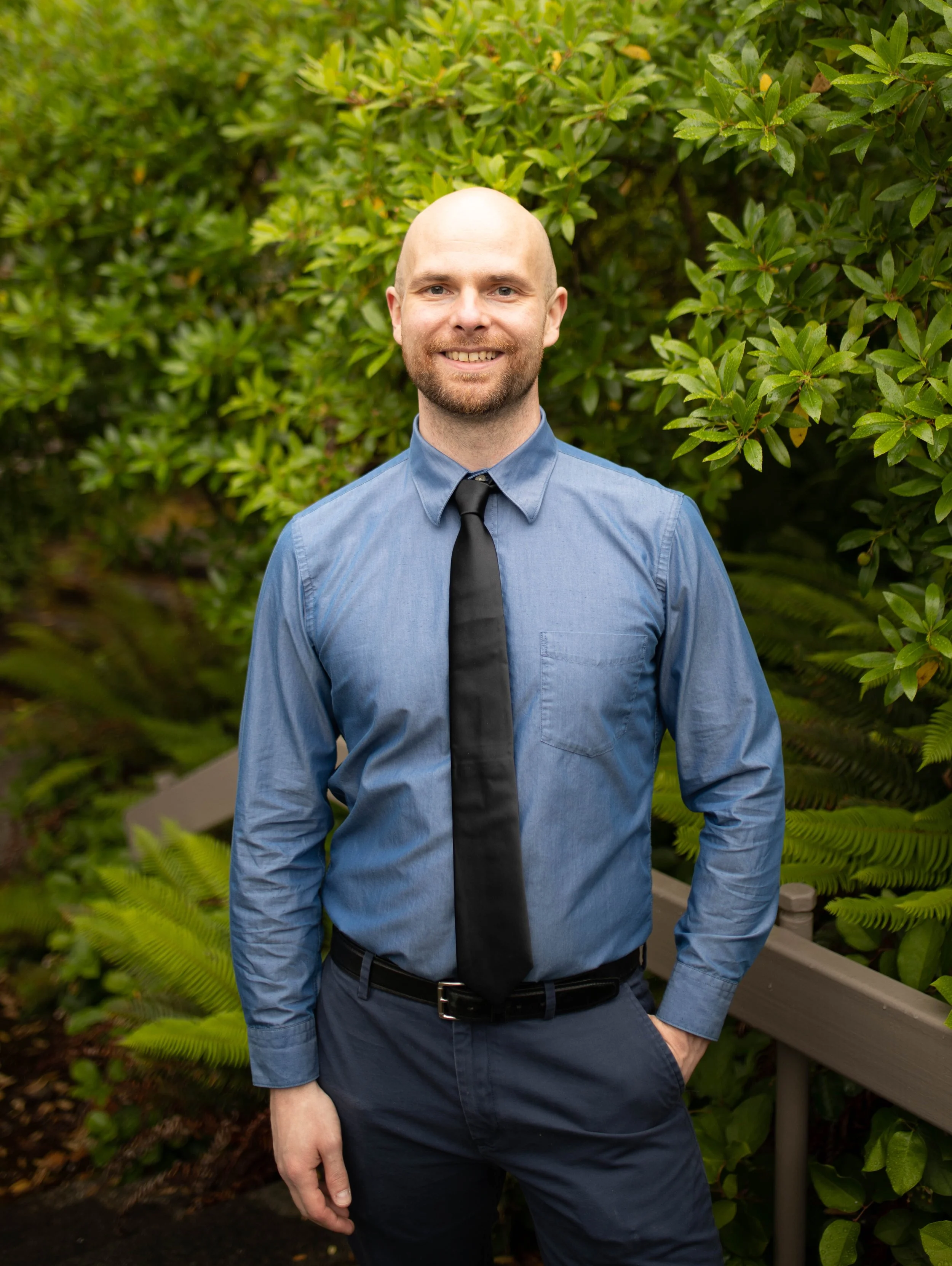 A man dressed in a blue shirt and black tie standing outdoors in front of green bushes with a small handrail behind him, smiling at the camera. Seattle professional head shot photography