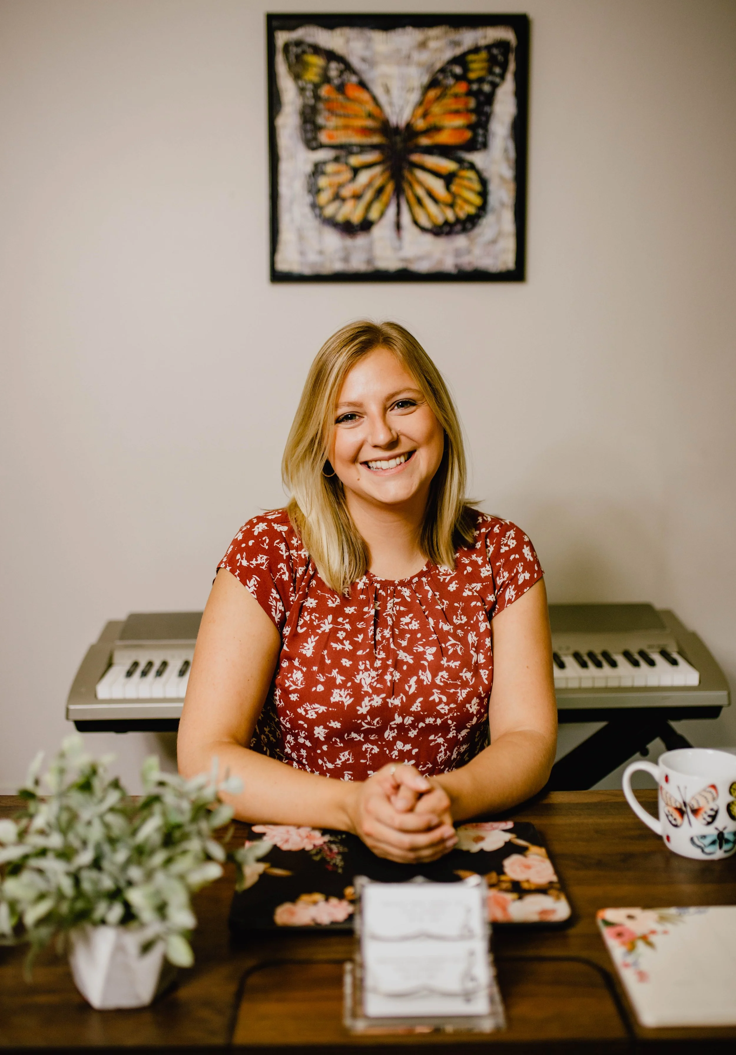 A woman with blonde hair smiling, sitting at a table with a floral tablecloth, a potted plant, a mug with butterfly designs, and a keyboard behind her. There is a butterfly picture hanging on the wall behind her. Seattle professional head shot photog