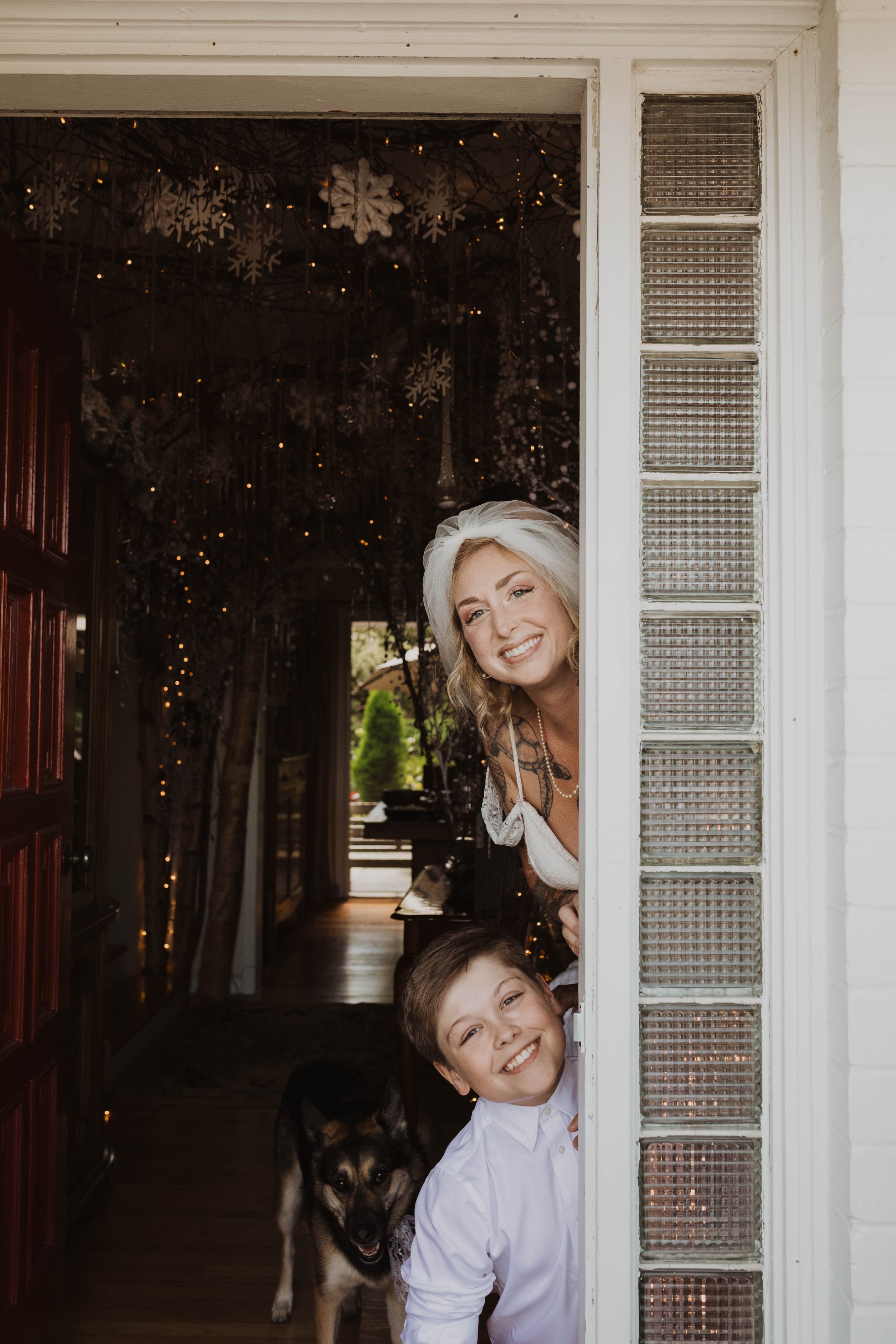 A woman, a boy, and a dog leaning out from a doorway, smiling at the camera. The woman has blonde hair and is wearing a white top, the boy has short brown hair and is wearing a white shirt, and the dog is a black and white husky.